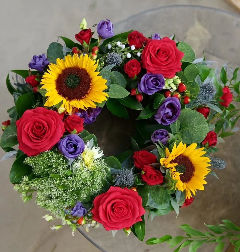 Colorful flower wreath with sunflowers, red roses, purple lisianthus, green foliage, and white accent flowers, on a gray surface background.