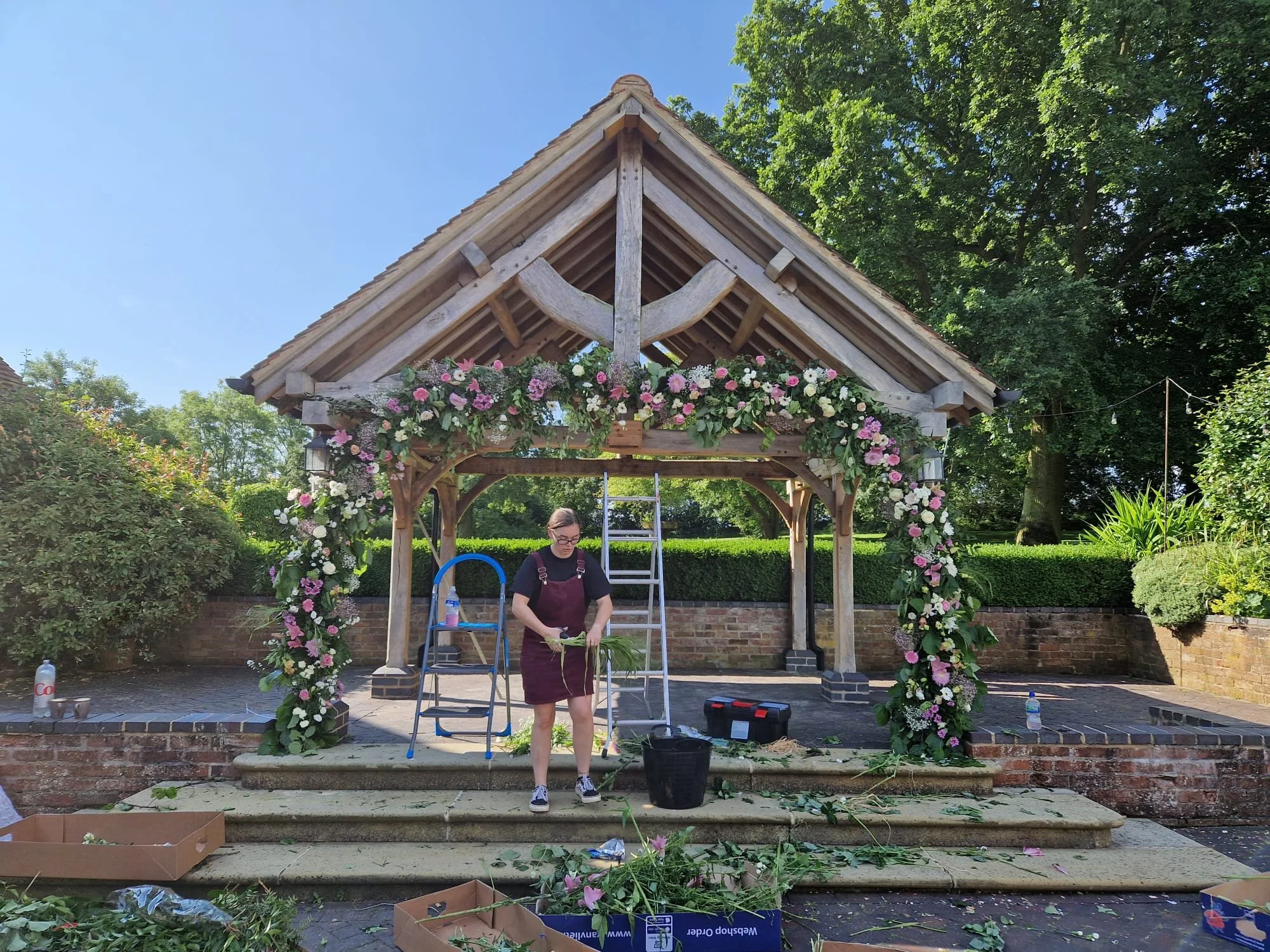 A woman decorating a wooden gazebo with pink and white flowers in an outdoor garden setting.