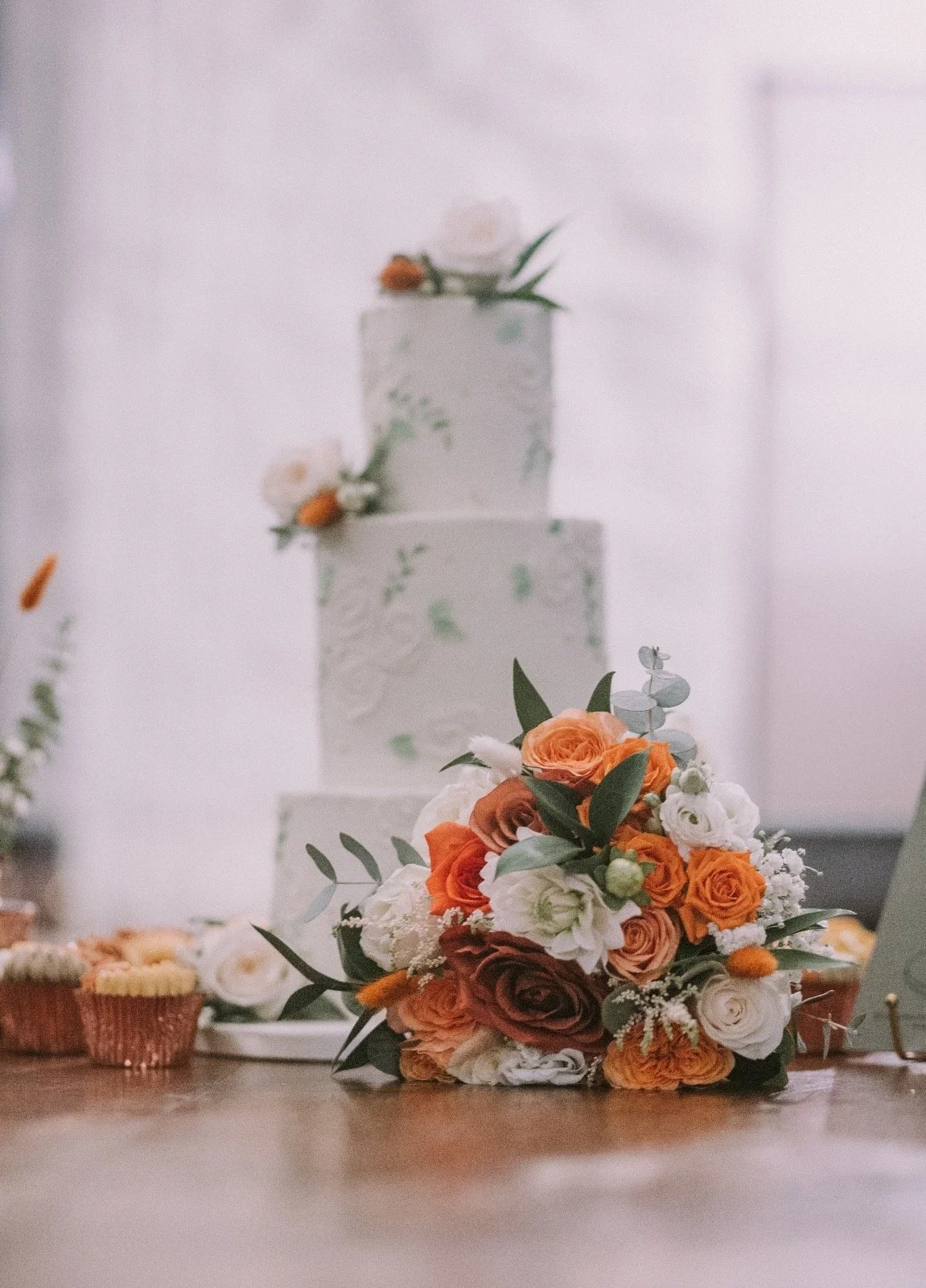 A wedding cake with a floral arrangement in front, displaying orange, white, and cream flowers, with a tiered white cake topped with similar flowers in the background.