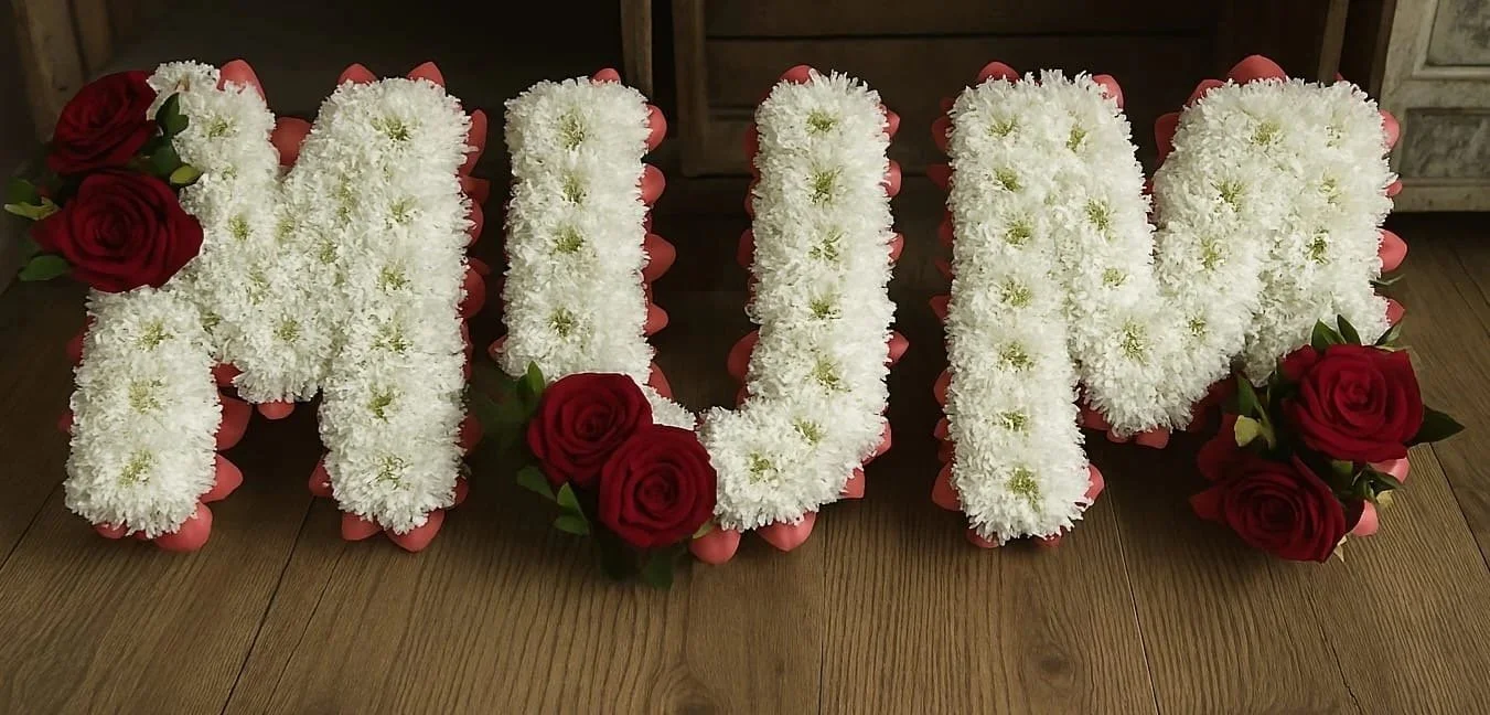 Flowers arranged to spell out 'MUM' with white flowers and red roses on a wooden floor.