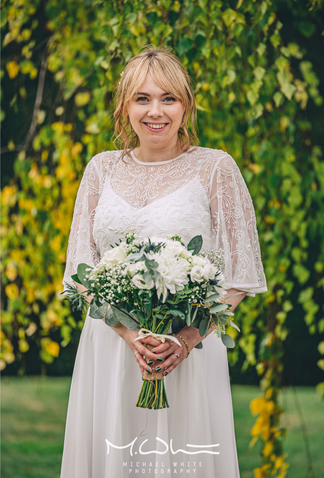 A woman in a wedding dress holding a bouquet of white flowers outdoors, smiling at the camera, with lush green foliage in the background.