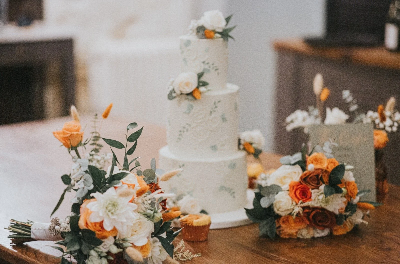 Wedding cake with white flowers and green leaves, floral bouquets with orange and white flowers, and cupcakes on a wooden table.
