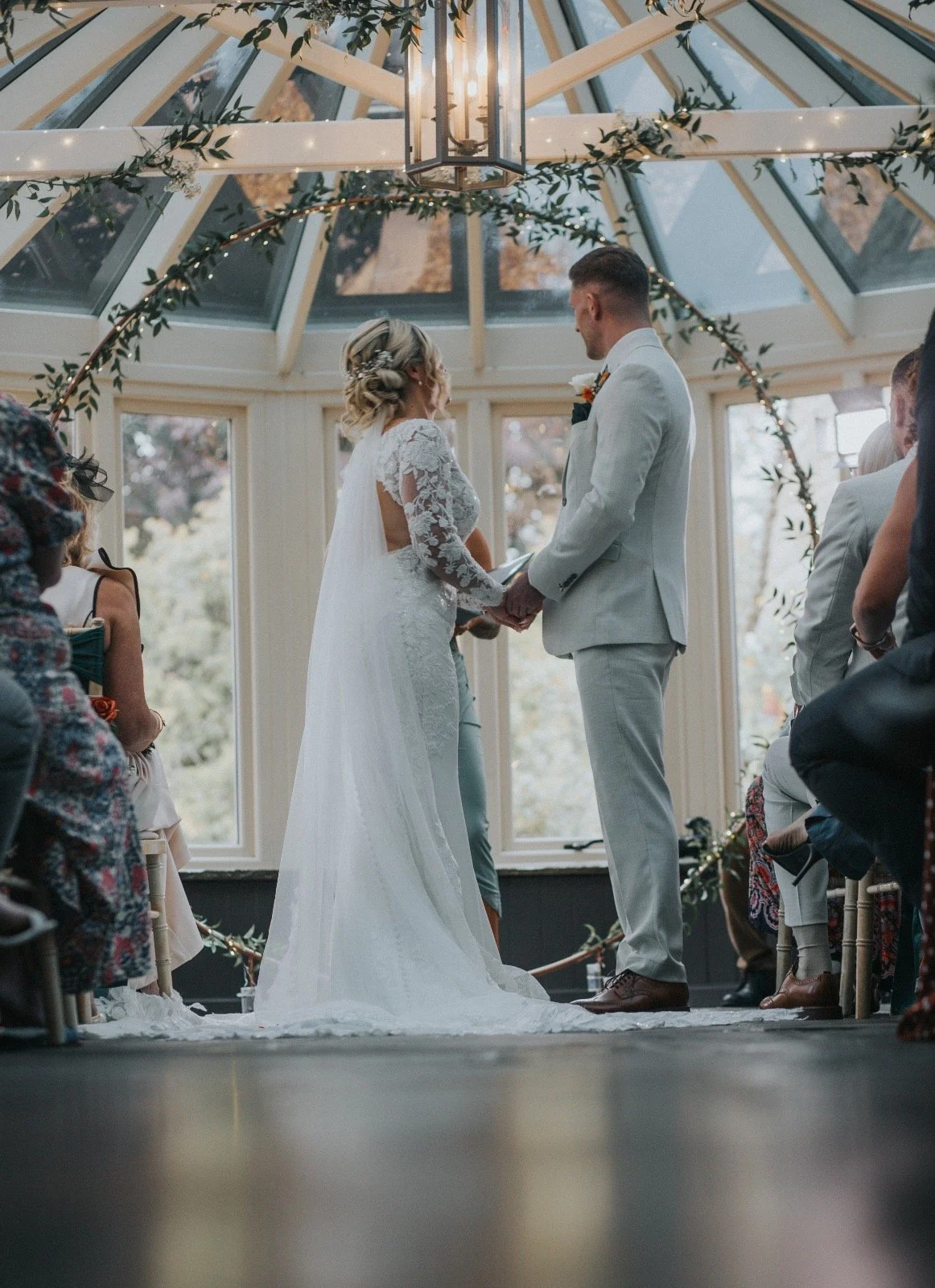 A bride and groom exchange vows during their wedding ceremony inside a bright, glass-ceilinged venue decorated with greenery and string lights.
