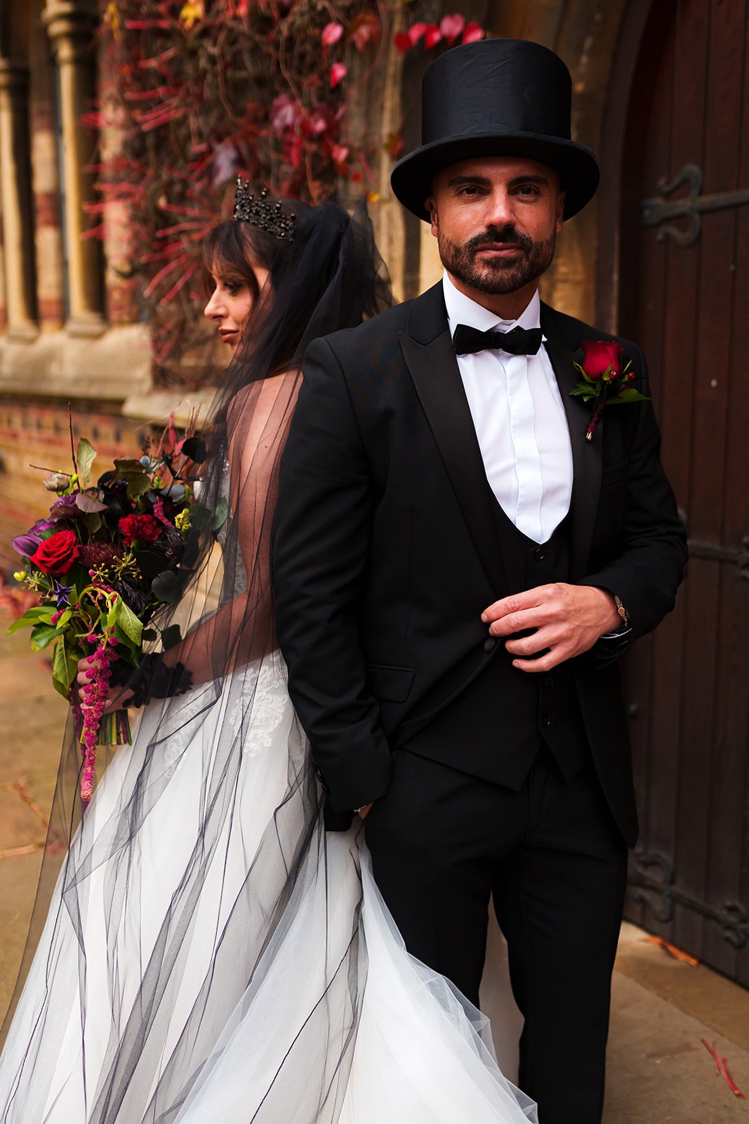 A groom dressed in a black tuxedo with a white shirt, black bow tie, and a red boutonniere, stands with his hand on his stomach, looking confidently at the camera. Behind him, the bride in a white wedding gown with black accents, a crown, and a black veil is facing sideways, holding a bouquet of colorful flowers, with a backdrop of a wooden door and autumn leaves.