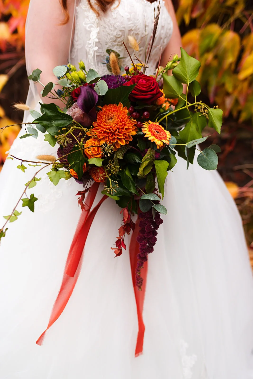 A bride in a white dress holding a cascading bouquet of orange, red, purple, and green flowers with green leaves and red ribbons, outdoors with colorful autumn foliage in the background.