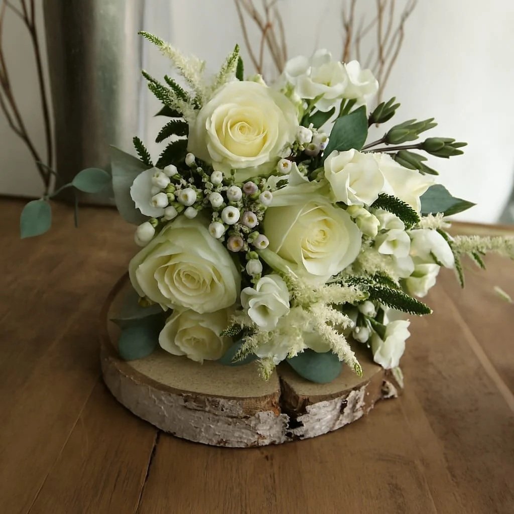 A bouquet of white roses, greenery, and small white flowers arranged on a wooden slice on a wooden table.