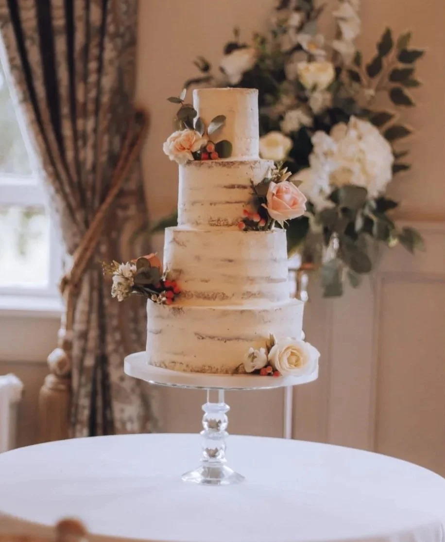 A four-tier wedding cake decorated with pink and white roses and greenery, placed on a glass cake stand in a decorated room with floral arrangements and curtains.
