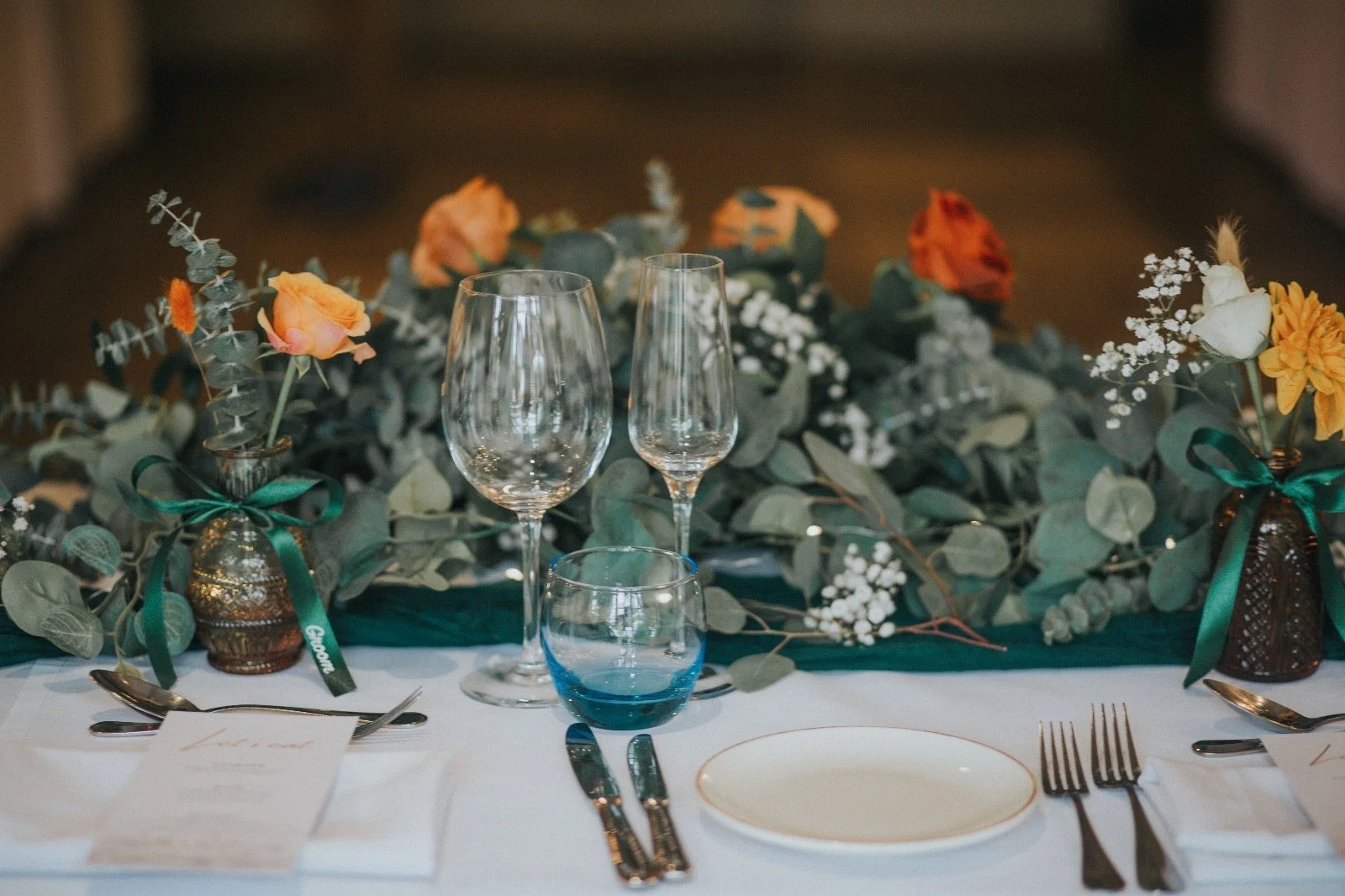 Elegant table setting with floral centerpiece, empty wine and champagne glasses, silverware, and candy-colored flowers in vases, likely for a formal event or celebration.