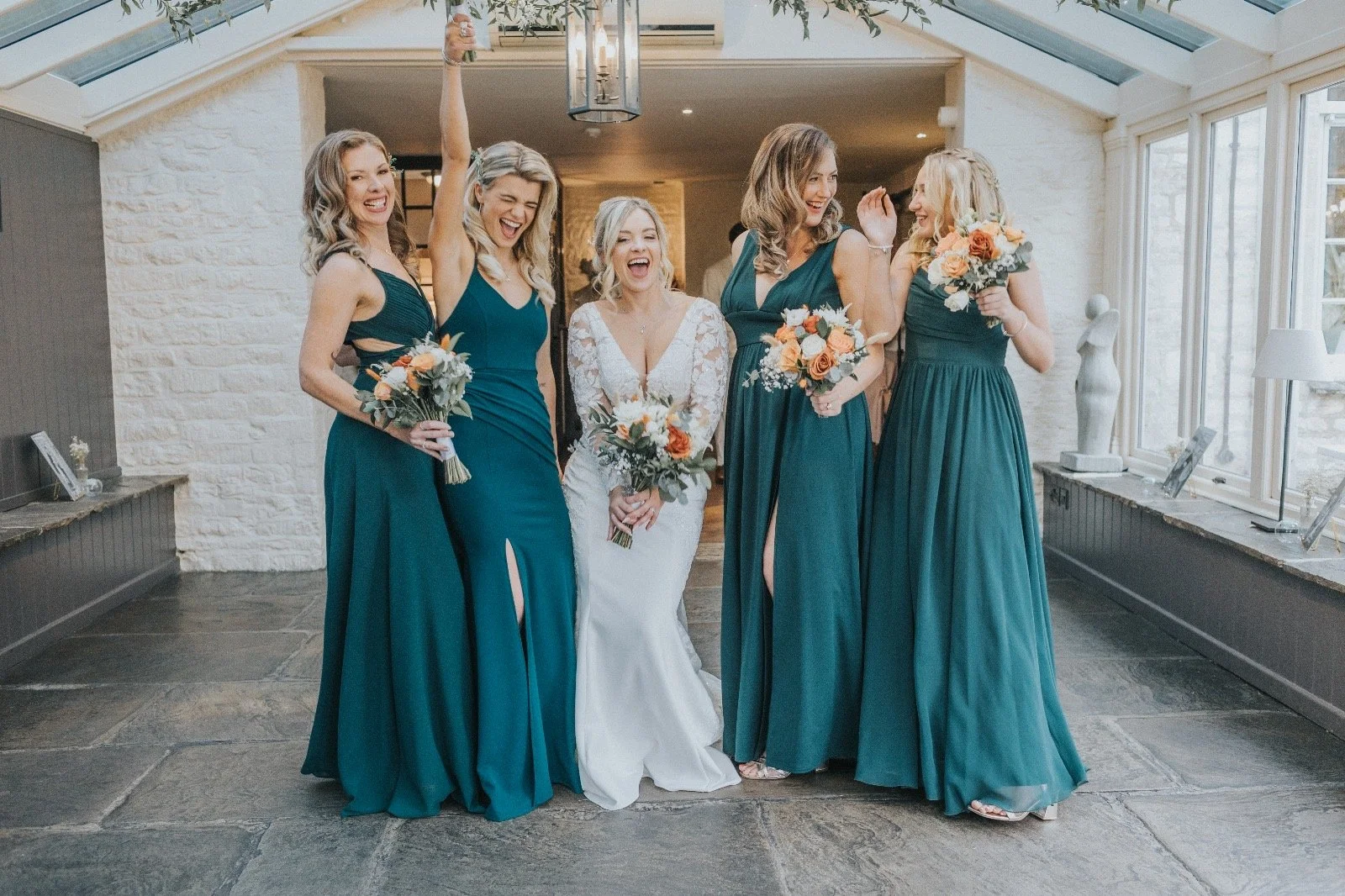 A bride in a white lace wedding dress and five bridesmaids in teal dresses, holding bouquets, celebrating indoors with large windows and a stone floor.