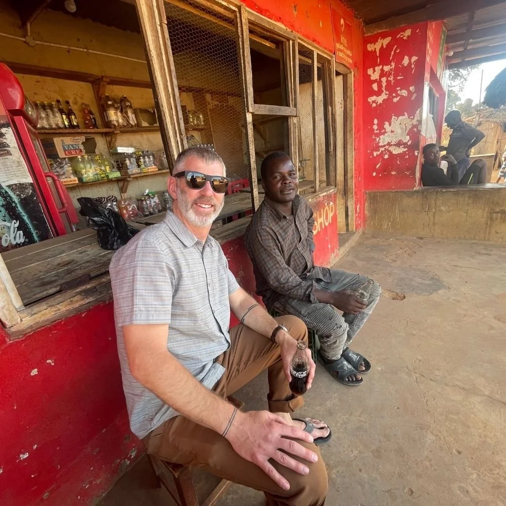 Two men sitting outside a small shop. One man is smiling, wearing sunglasses, a light shirt, and holding a bottle of soda. The other man is dressed in a checkered shirt and striped pants. Behind them are shelves with bottles and jars, and a red Coca-Cola refrigerator.