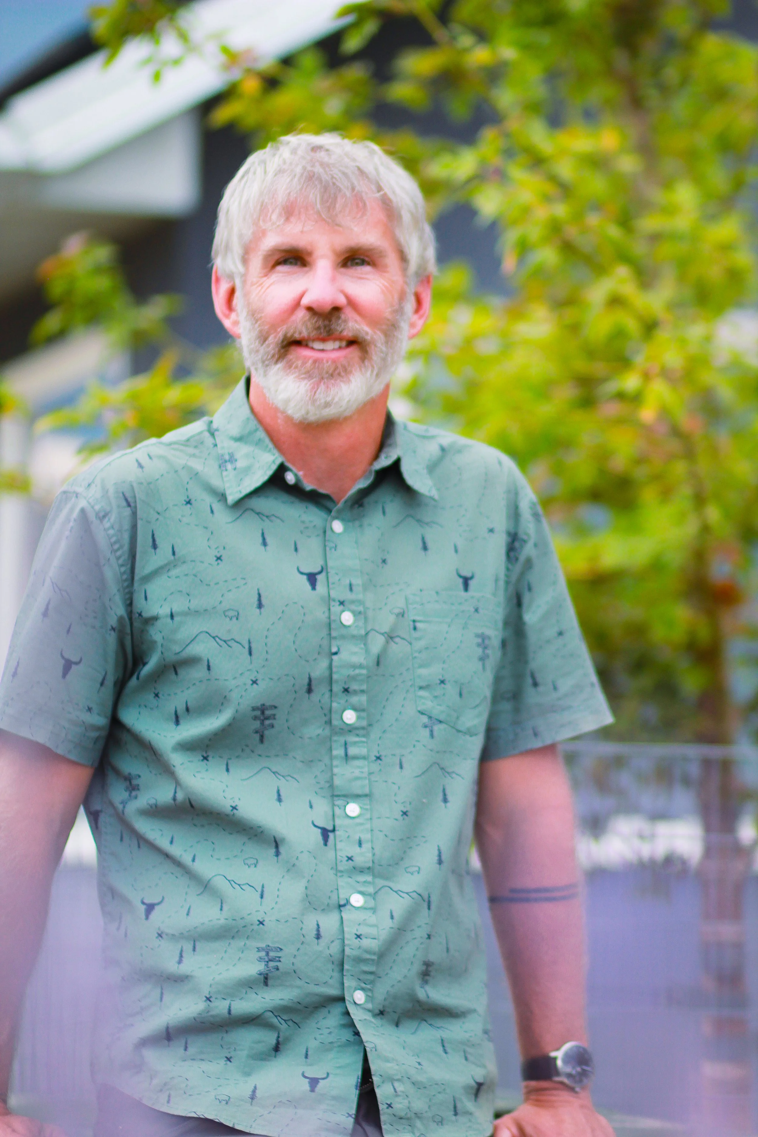 Close-up of a smiling man with gray hair and beard, wearing a patterned short-sleeve shirt, standing outdoors with green trees in the background.