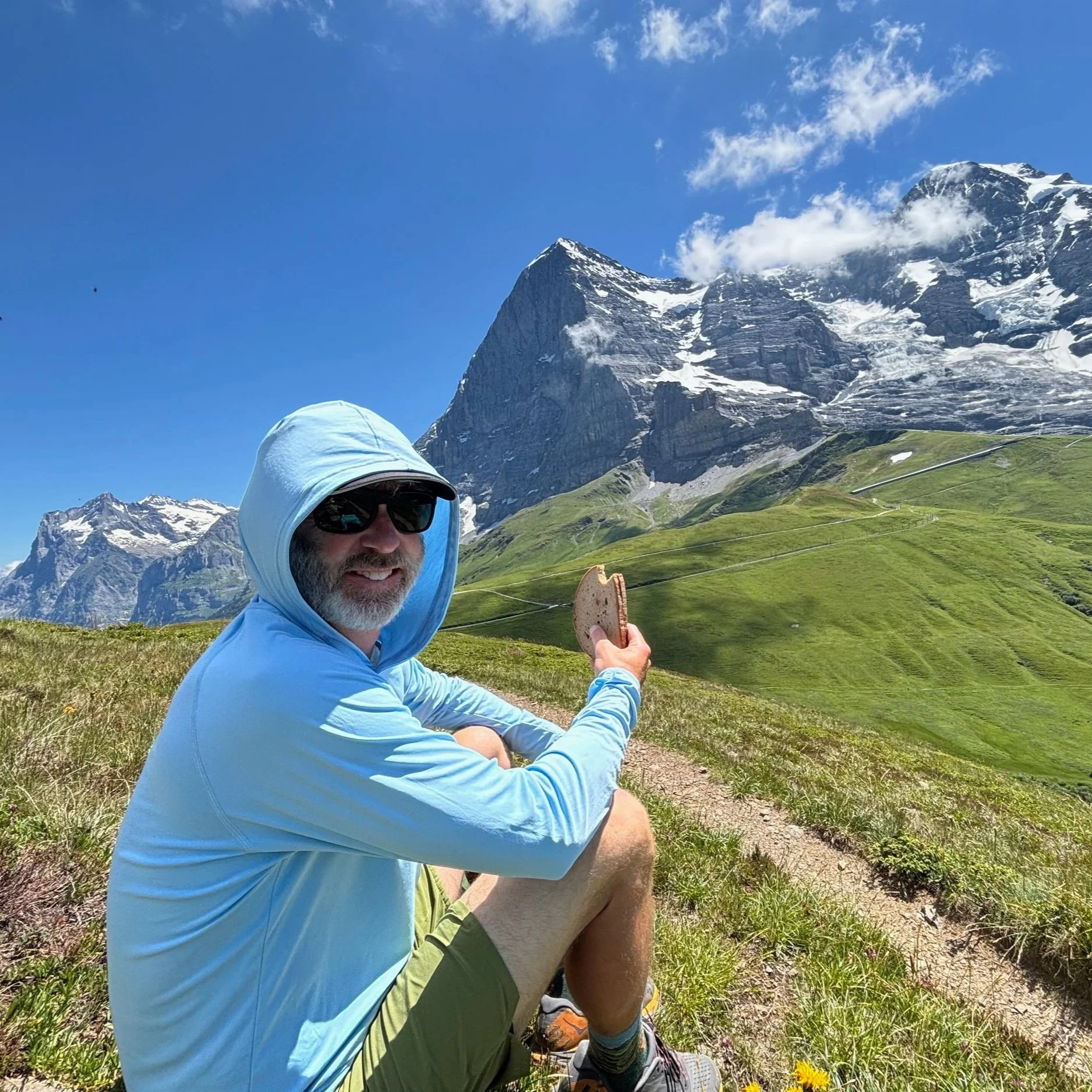 A person in outdoor clothing, including a light blue jacket and sunglasses, sitting on a grassy mountain path with snow-capped peaks and blue sky in the background, holding a snack.