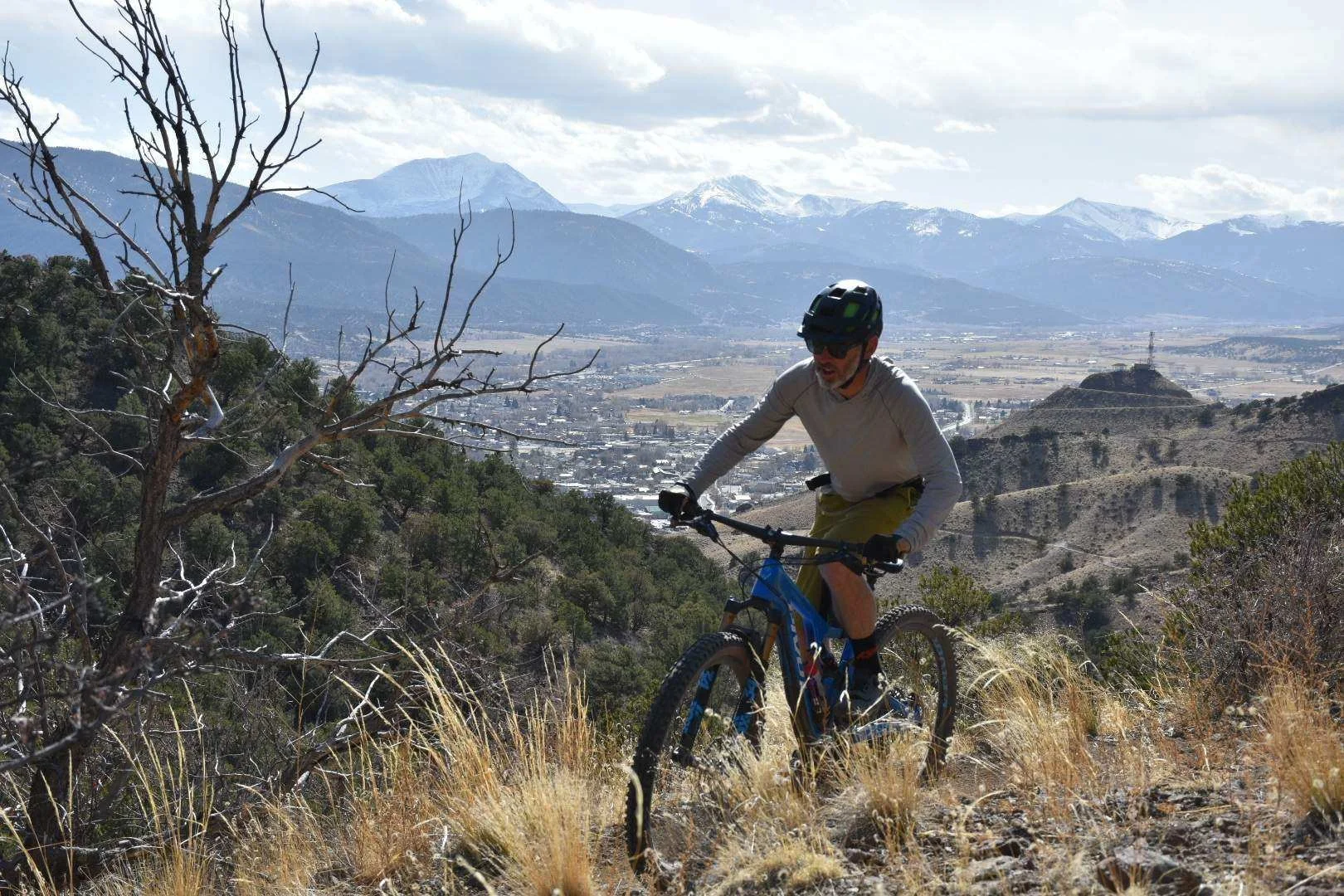 A man mountain biking on a trail in a dry, hilly landscape with mountains in the background, some with snow, and a partly cloudy sky.