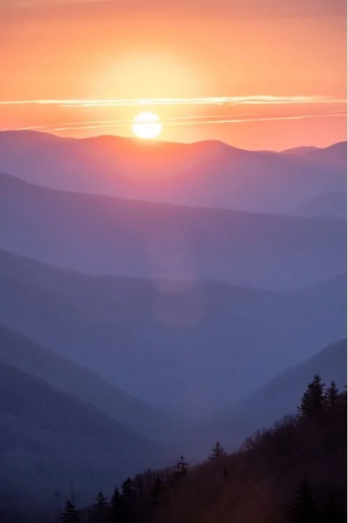 Sun setting over layered mountains at sunset with pink, orange, and purple hues in the sky.
