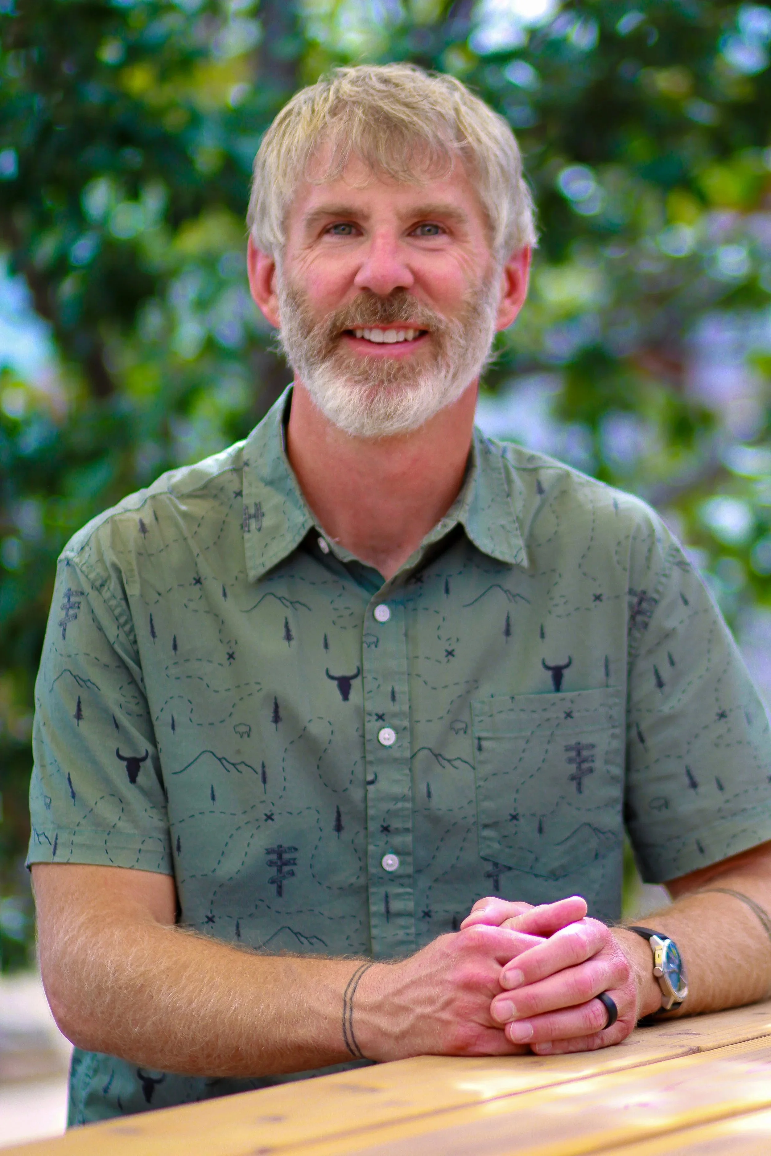 A man with gray hair and a beard sitting at a table outdoors, smiling, wearing a patterned shirt, with trees in the background.