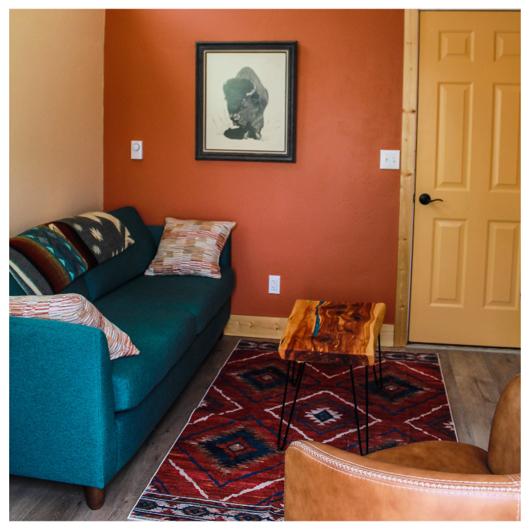 A cozy living room corner with a teal sofa decorated with patterned pillows, a small wooden side table with black hairpin legs, a patterned area rug, a brown leather chair, and a framed abstract black-and-white artwork on an orange accent wall next to a tan door.