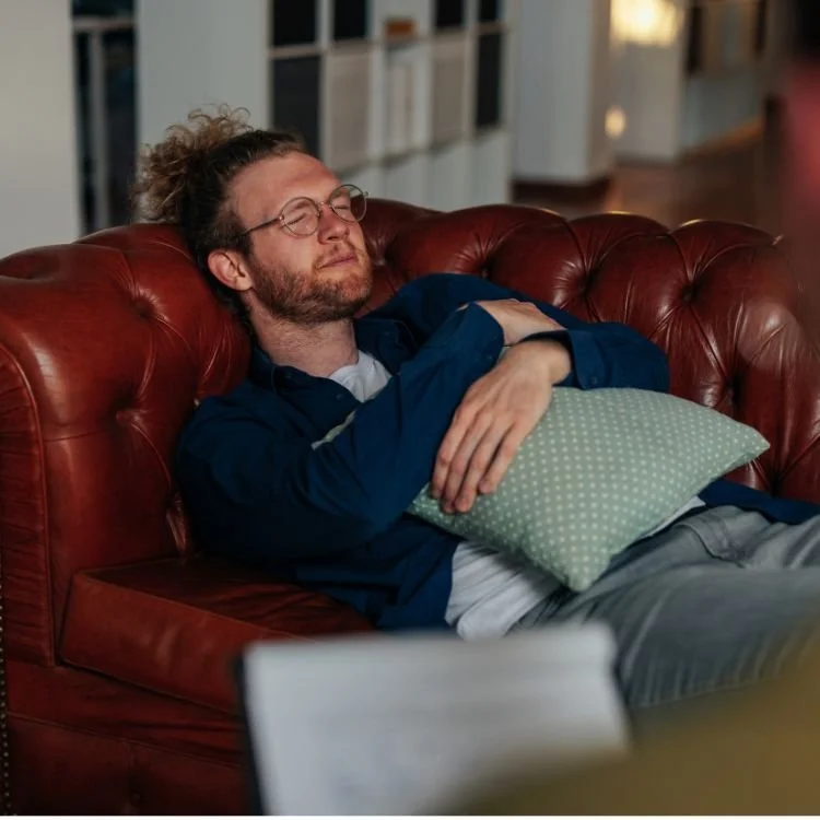 Man with glasses and curly hair sleeping on a red leather couch, holding a green pillow.