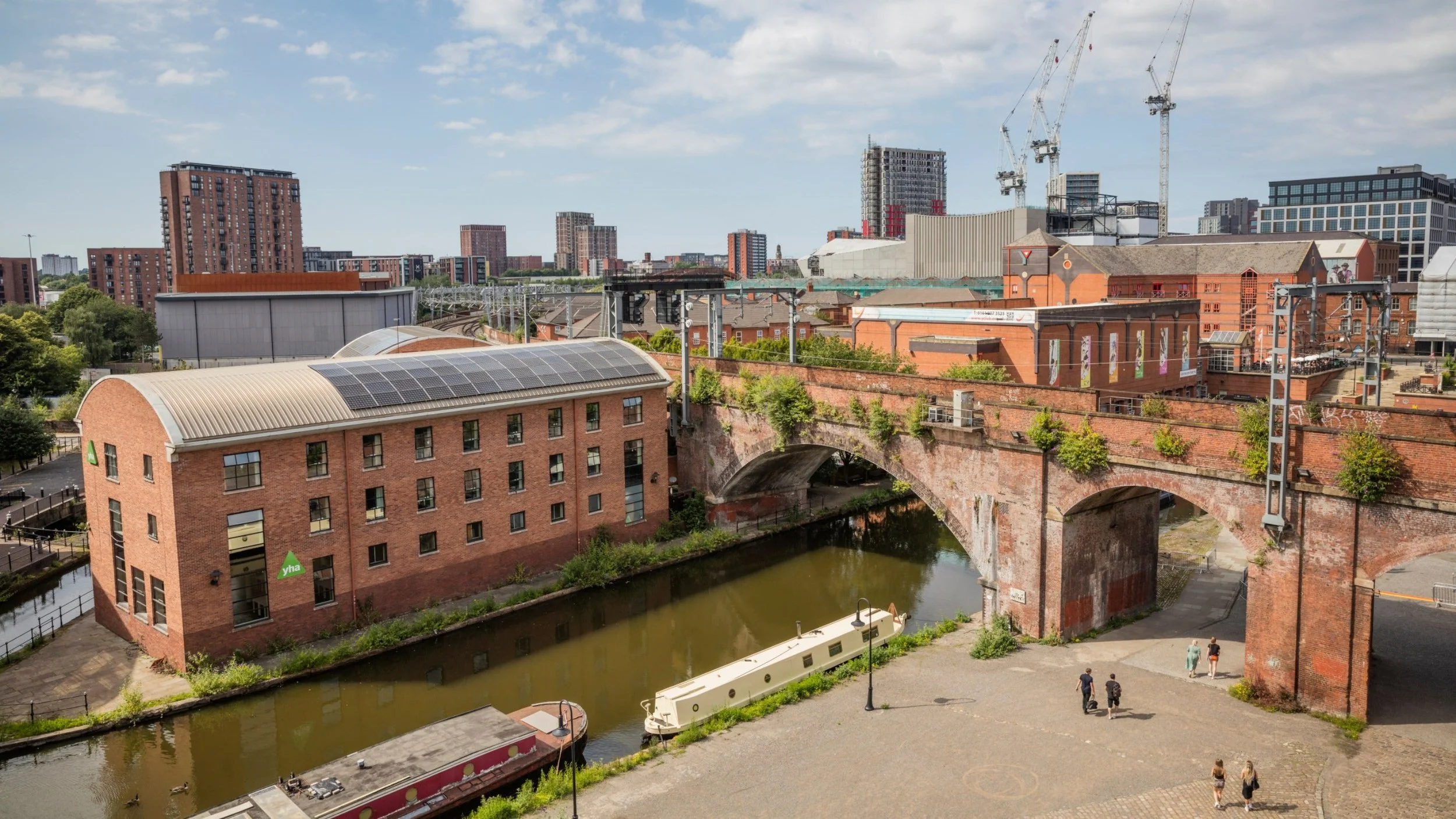 Cityscape with brick and modern buildings, a bridge over a canal, boats, and a few people walking on the street.