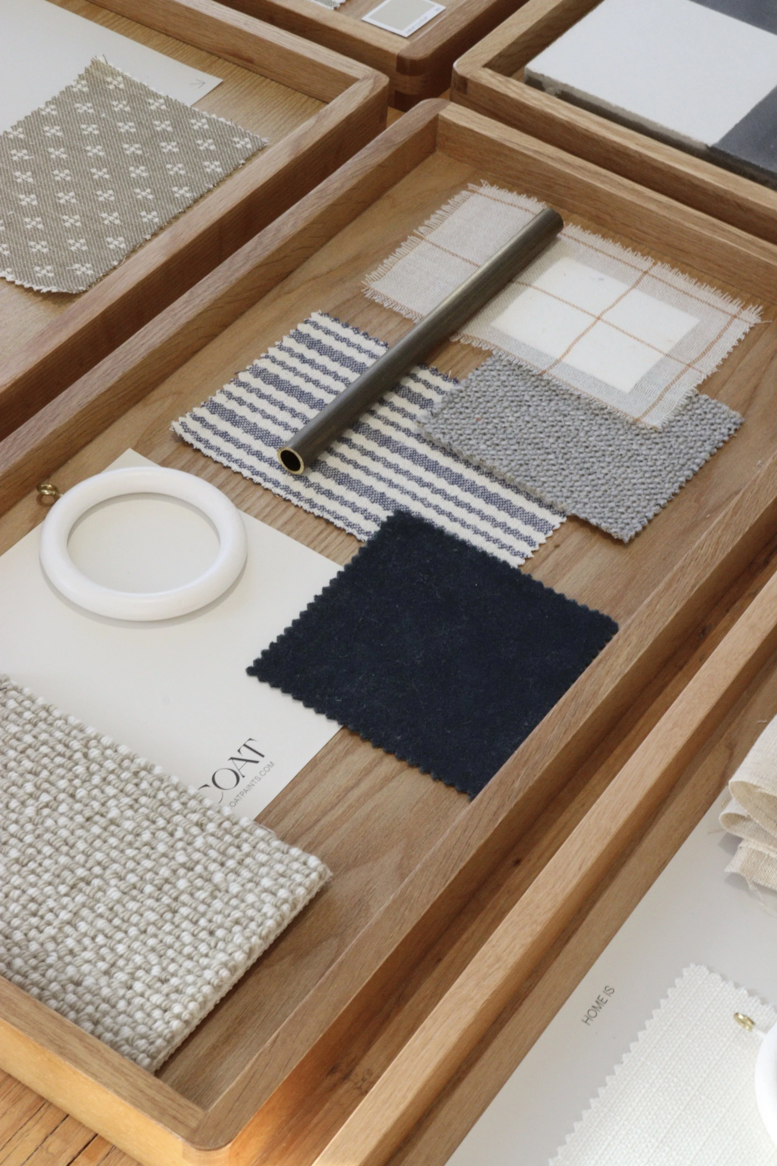 A wooden display tray containing fabric samples in neutral tones, including woven, striped, and checkered patterns, with a metal rod and a small white ring.