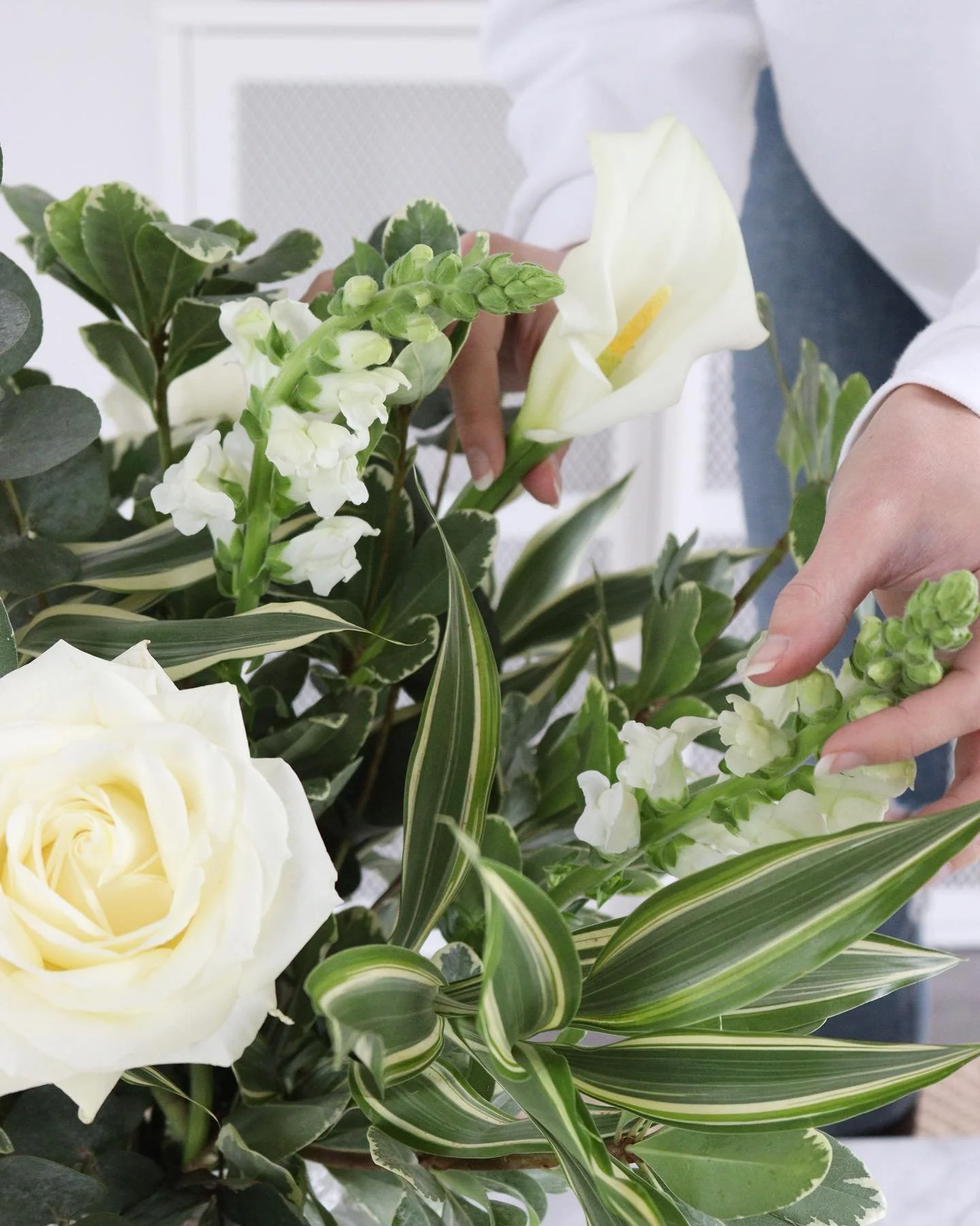 Person arranging white flowers, including roses and lilies, in a bouquet with green leaves.