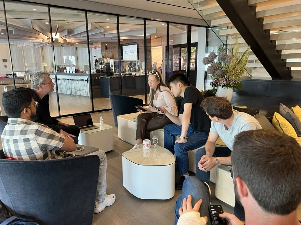 A group of six people sitting and talking in a modern waiting area with a glass partition behind them, and a coffee station visible in the background.