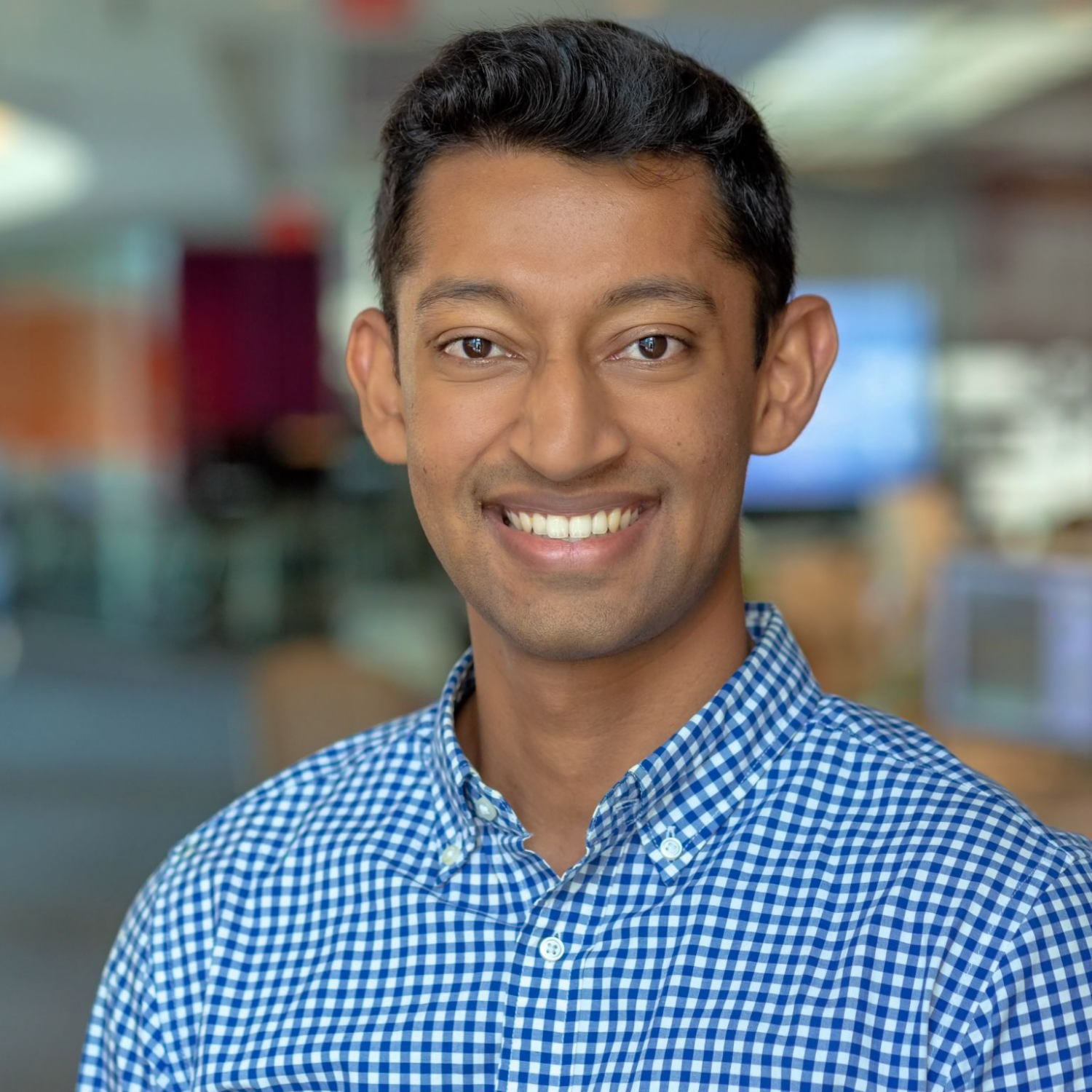 A smiling man with short black hair, wearing a blue and white checkered shirt, posing in an indoor setting with a blurred background.