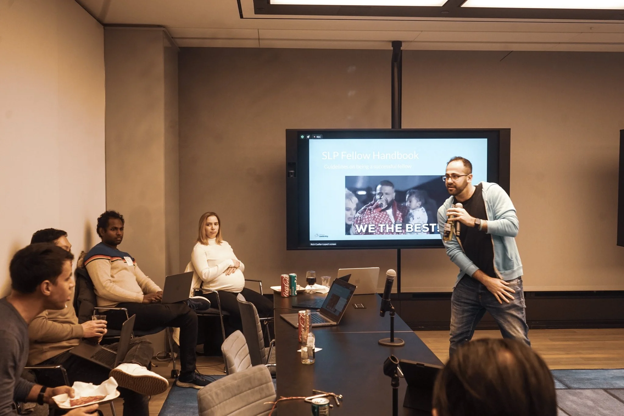 A group of people attending a presentation in a conference room. A man is speaking into a microphone in front of a large screen displaying a slide titled 'SLP Fellow Handbook.' Others are seated, some with laptops, and a pregnant woman is sitting with her hands resting on her belly.
