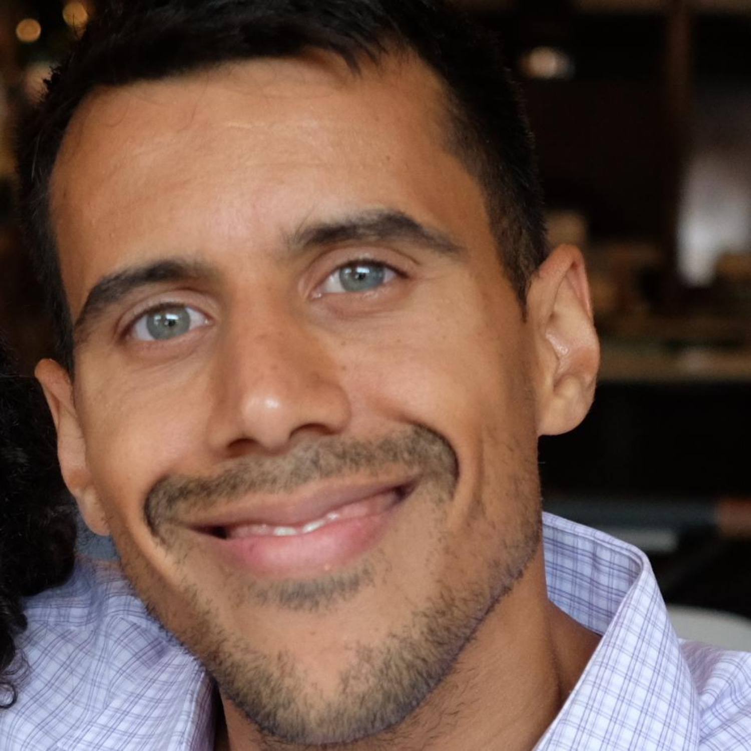 A smiling man with hazel eyes, dark hair, and a light beard wearing a checkered shirt.