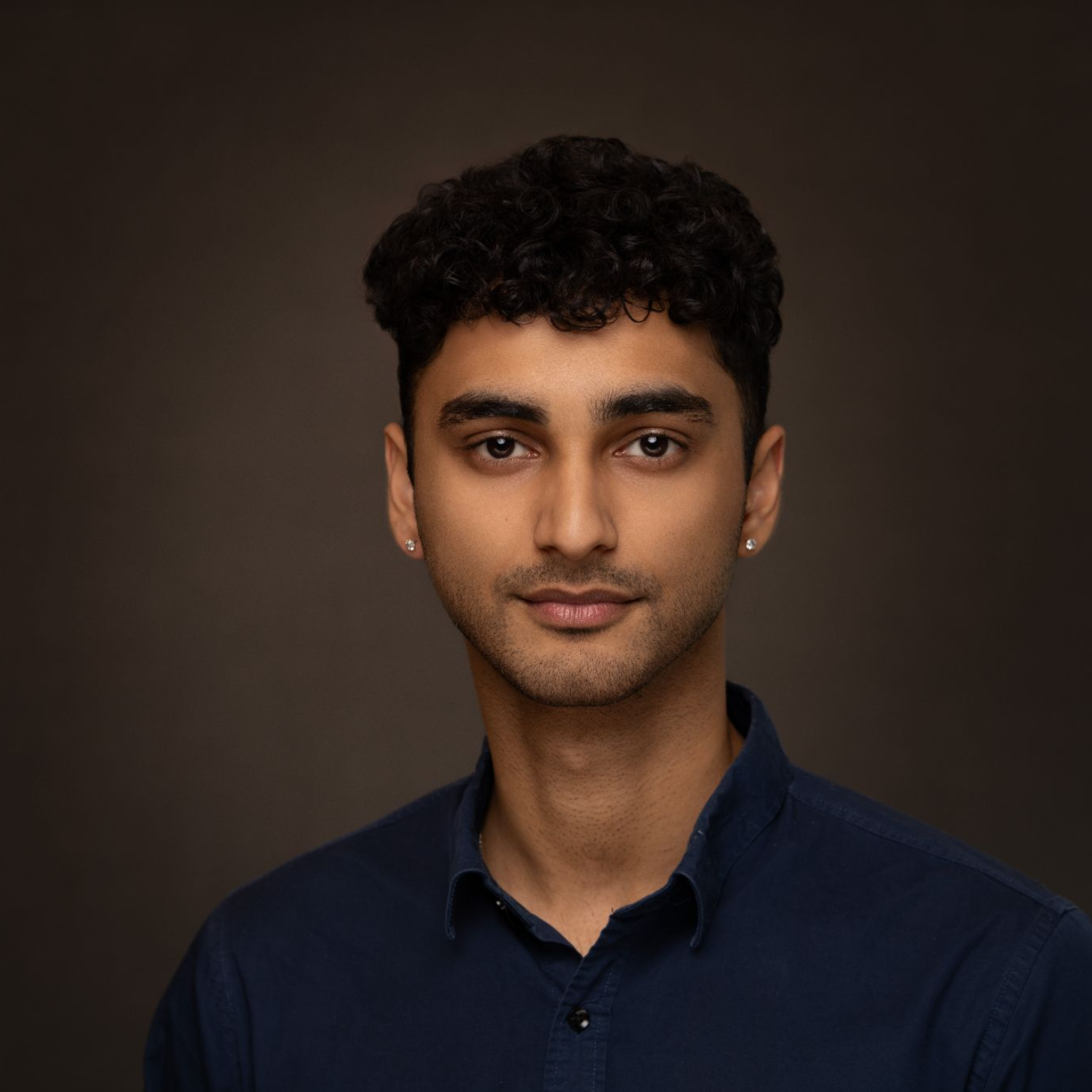 Portrait of a young man with dark curly hair, brown eyes, wearing earrings and a navy blue collared shirt, against a dark background.