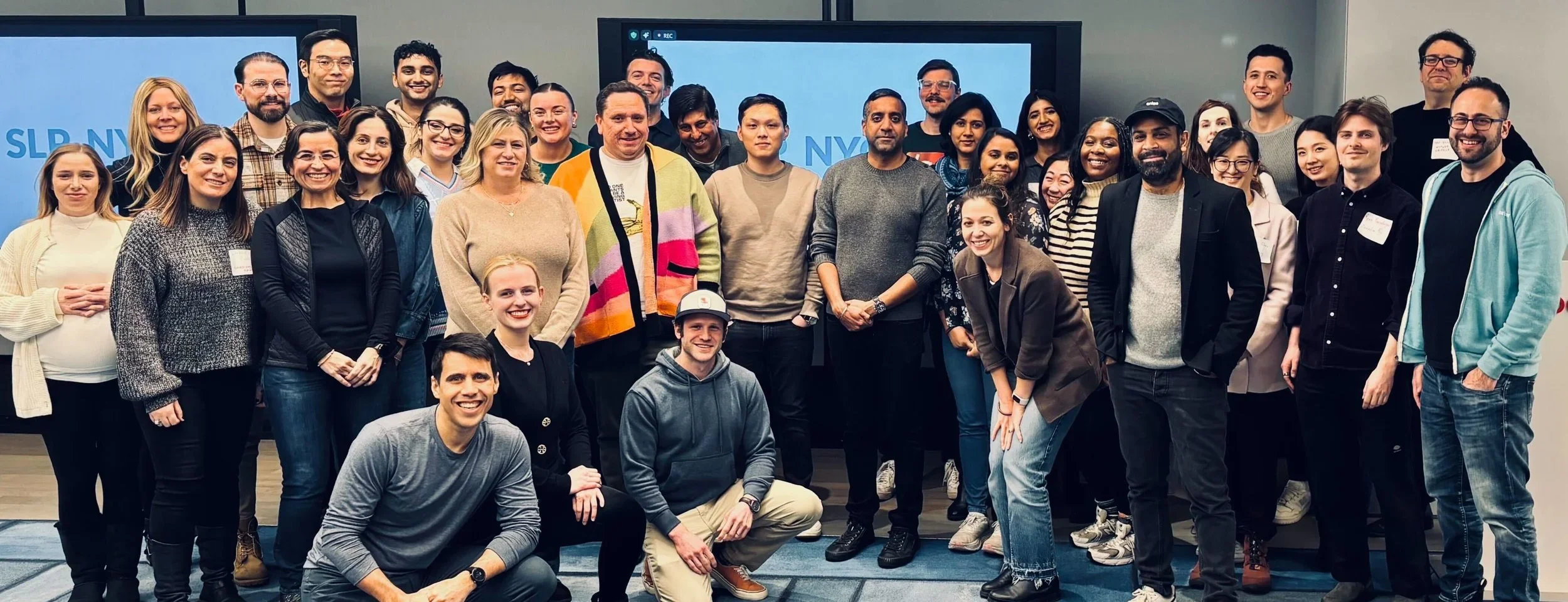 Group of diverse people in a conference room, several standing behind and some kneeling in front, smiling for a group photo.