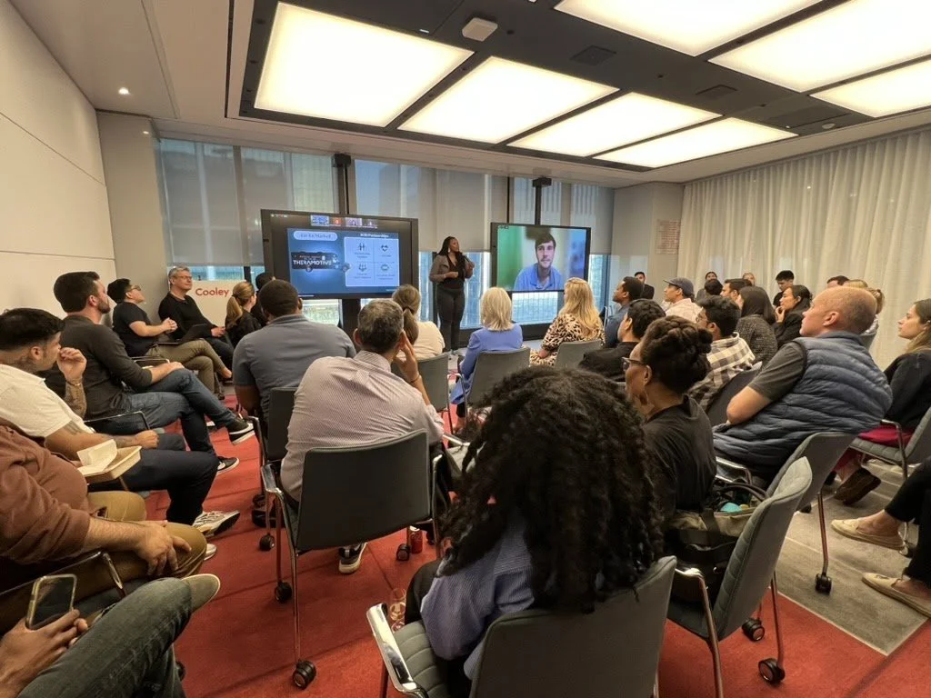 A conference room filled with people attending a presentation, with two large screens at the front, one displaying a woman speaking, and the other showing a man in a video call.