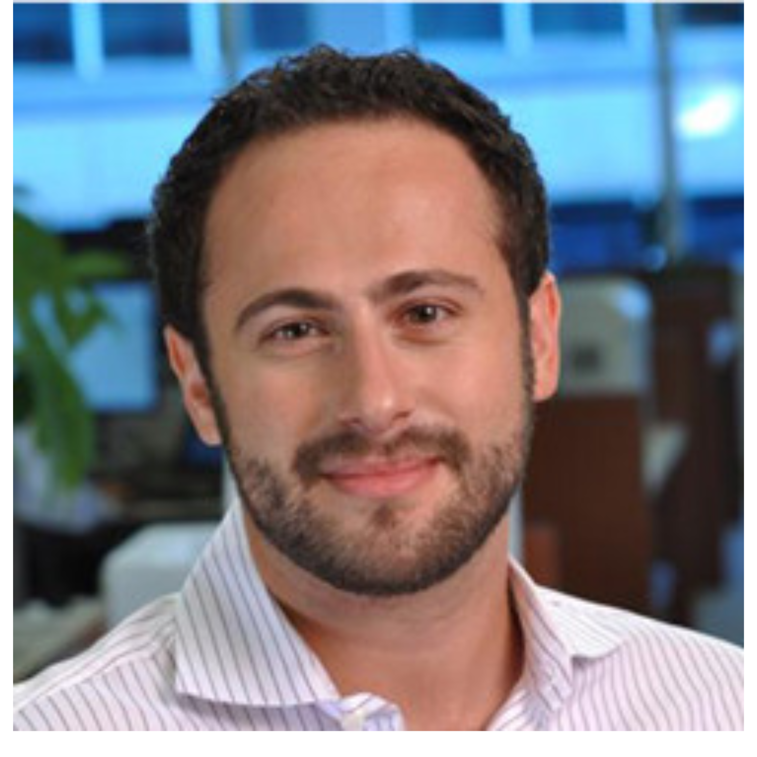 Headshot of a man with dark hair and beard, wearing a collared shirt, smiling in an office environment.