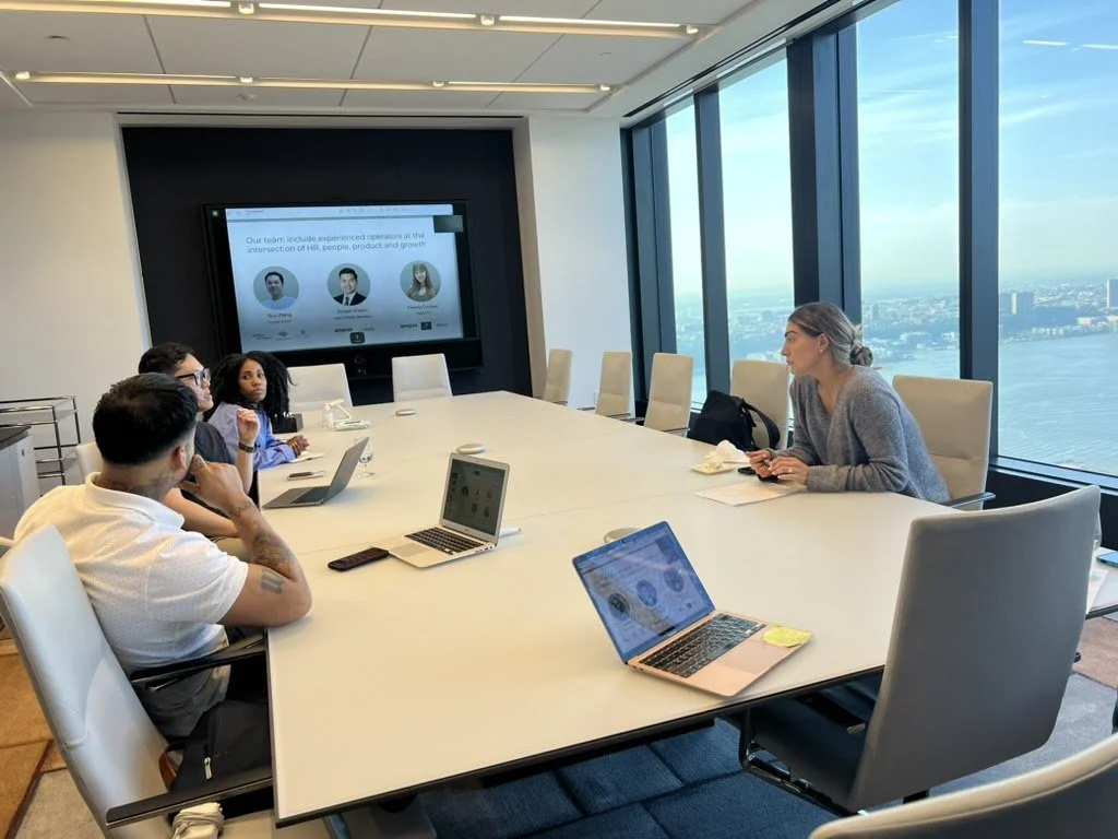 Business meeting in a high-rise conference room with large windows showing a city and river view. Five people are seated around a long table with laptops and notebooks, and a presentation screen displays information about a team.