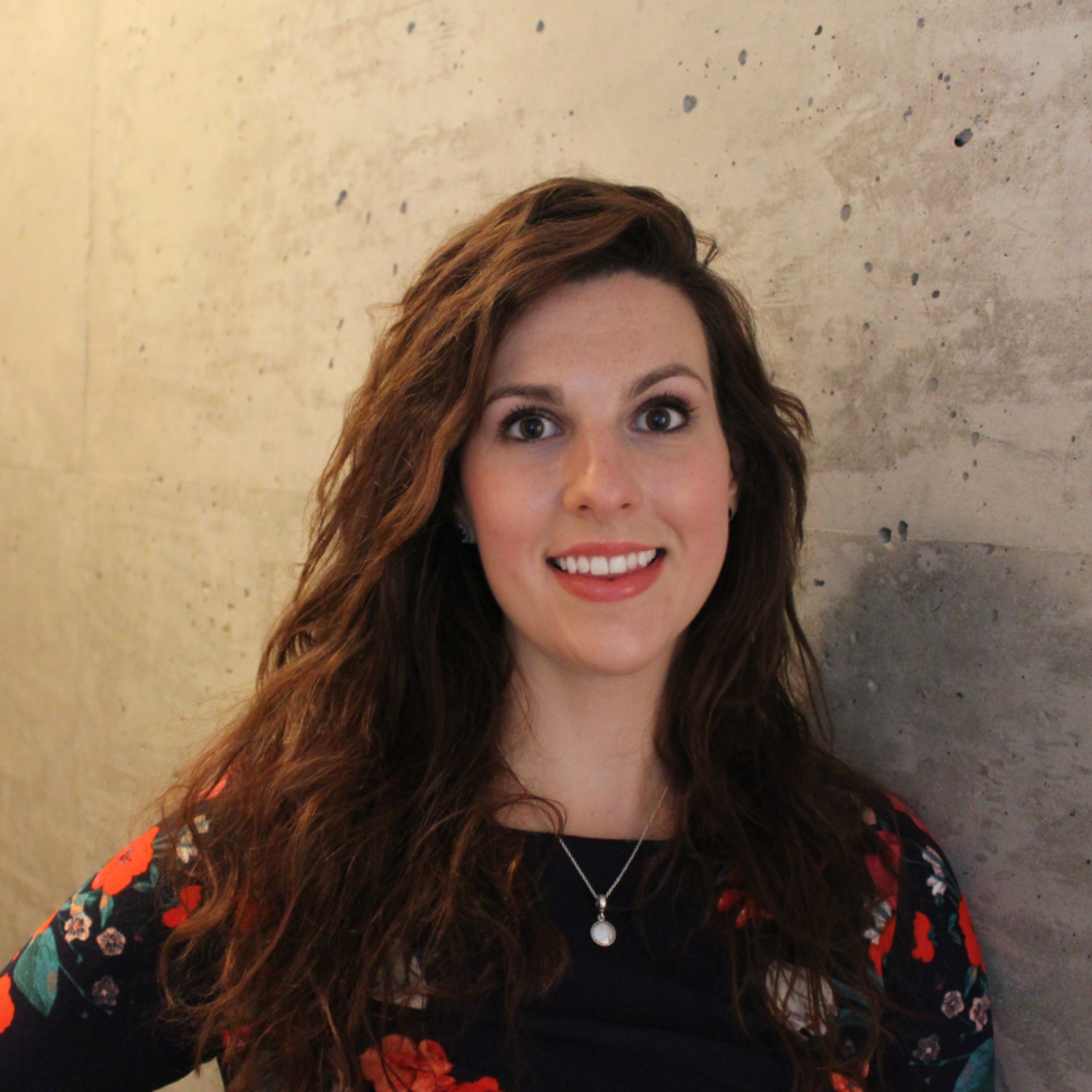 A woman with long wavy brown hair, wearing a black top with a colorful floral pattern, and a silver necklace, smiling in front of a grey textured wall.