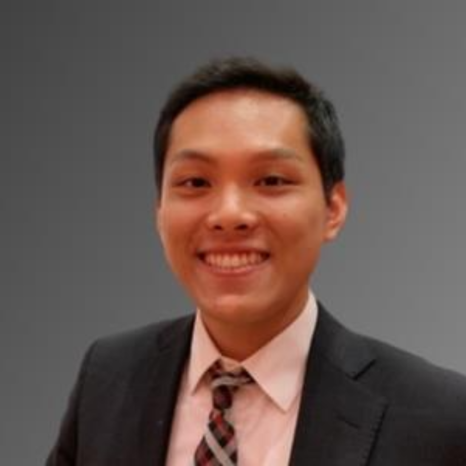 A young man with short dark hair wearing a suit, dress shirt, and patterned tie, smiling against a plain background.