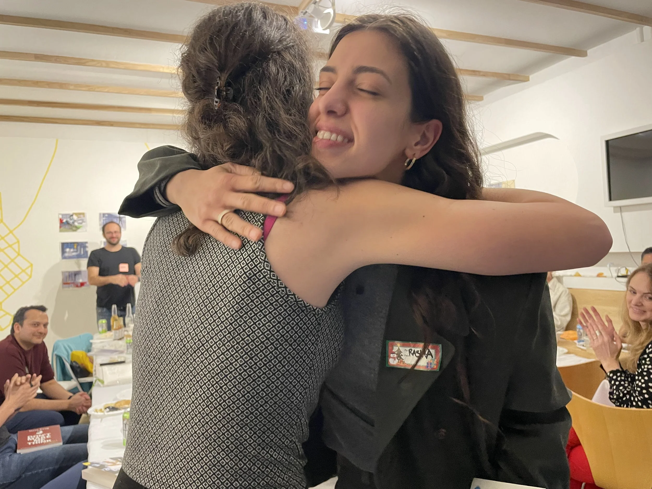 Two women are hugging each other tightly, one with her eyes closed smiling, at a social gathering. There are several people sitting at a table, clapping and smiling in the background.