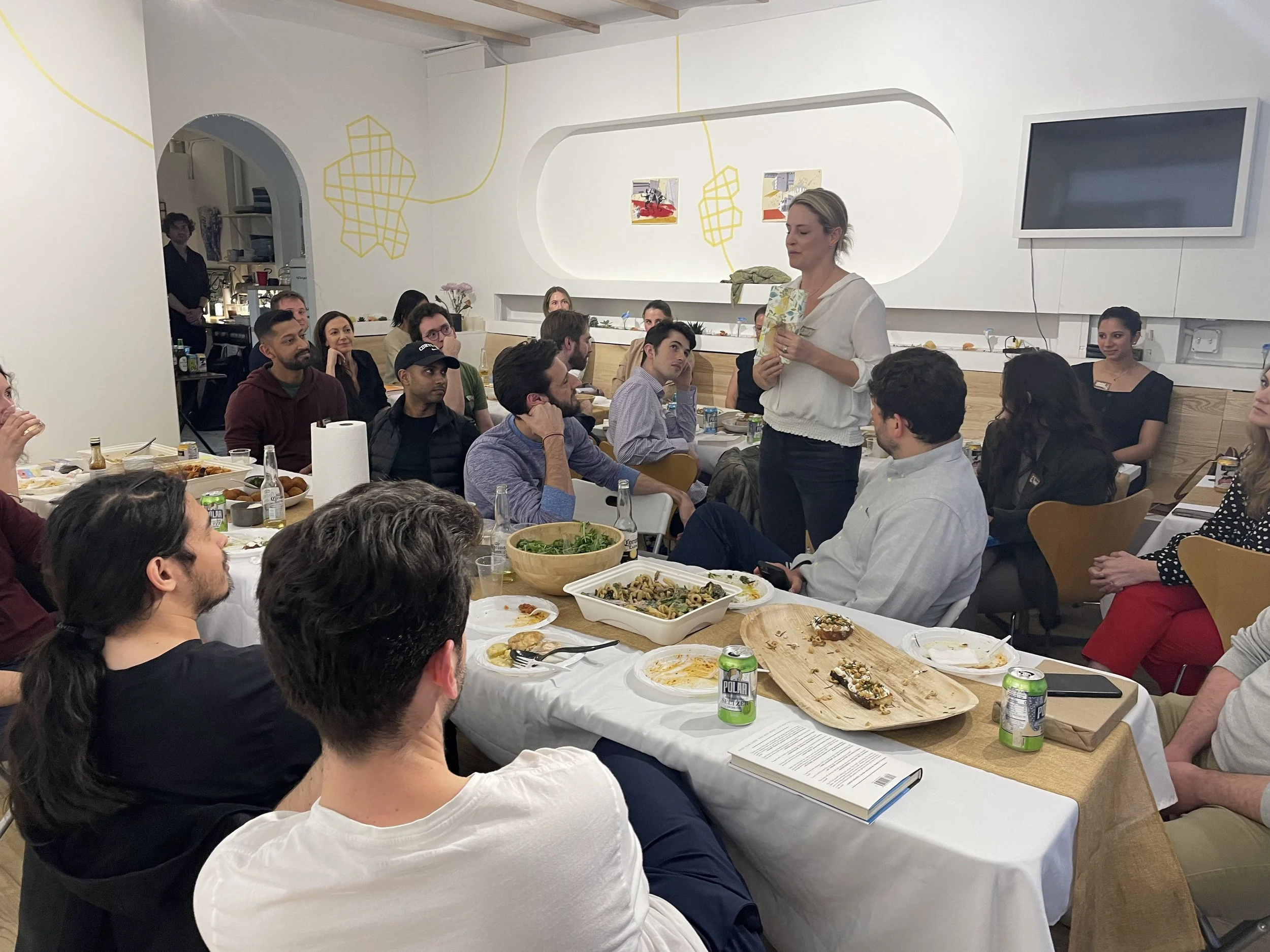 Group of people attending a presentation in a restaurant or cafe, with a woman standing and speaking to the audience, surrounded by tables with food and drinks.
