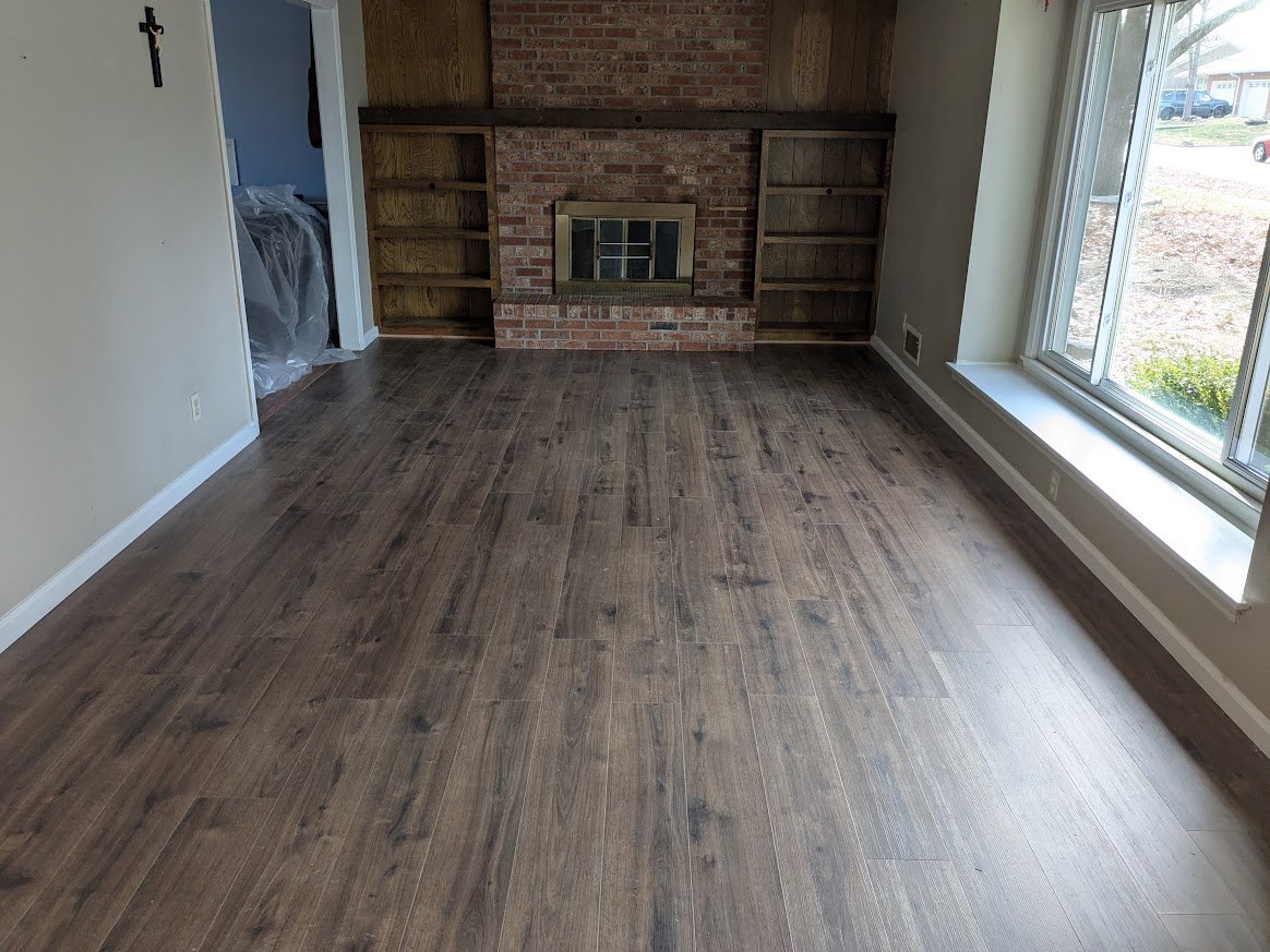 Empty living room with wood flooring, brick fireplace, built-in wooden shelves, large window letting in natural light, and a cross on the wall in the adjacent room.