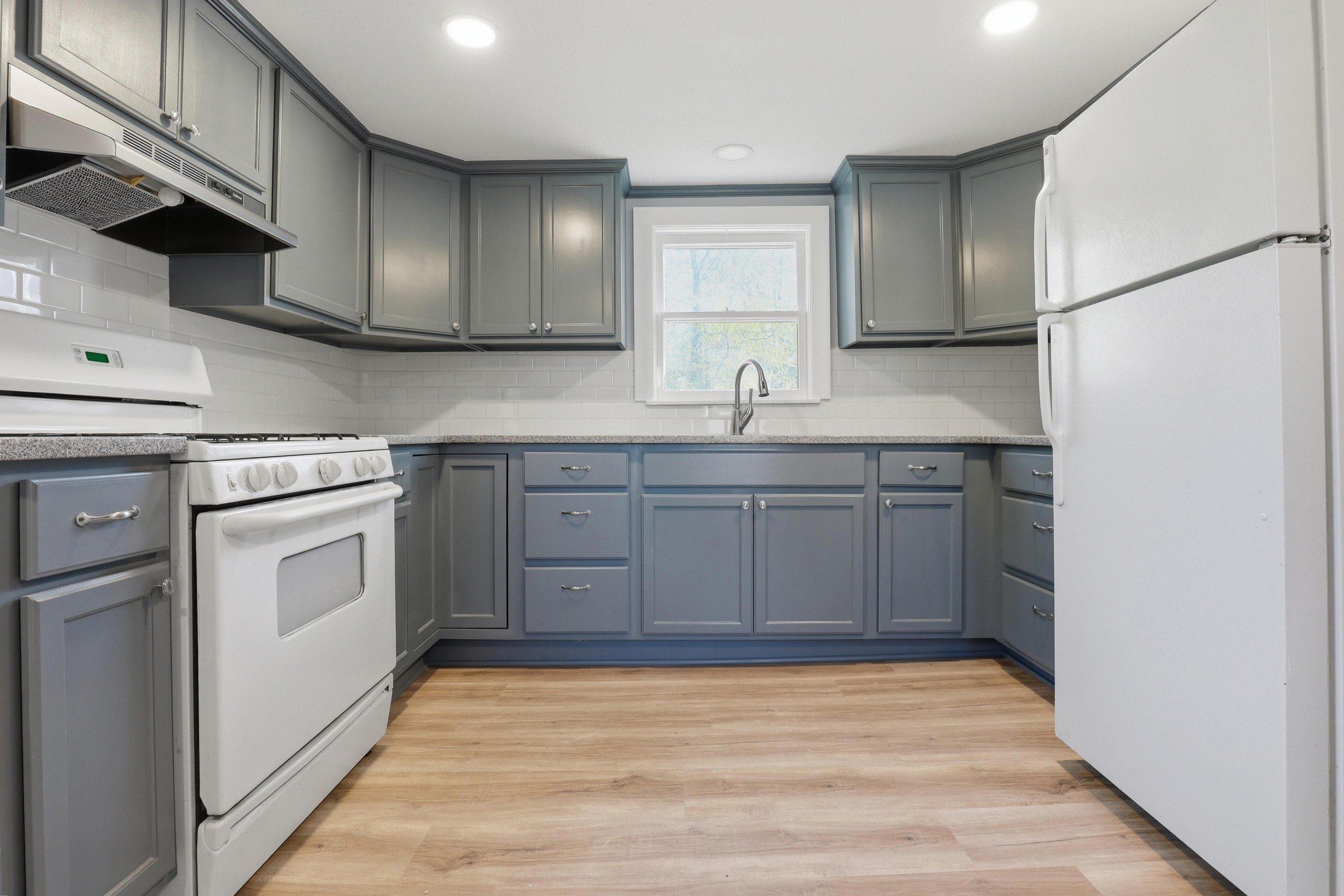 A kitchen with light gray cabinets, white appliances, a window above the sink, white subway tile backsplash, and wood flooring.