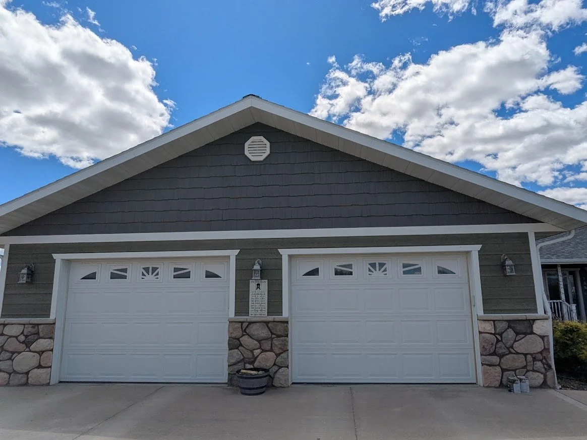 Front view of a house with two white garage doors, stone accents, and a dark gray exterior. The sky is partly cloudy with blue skies.