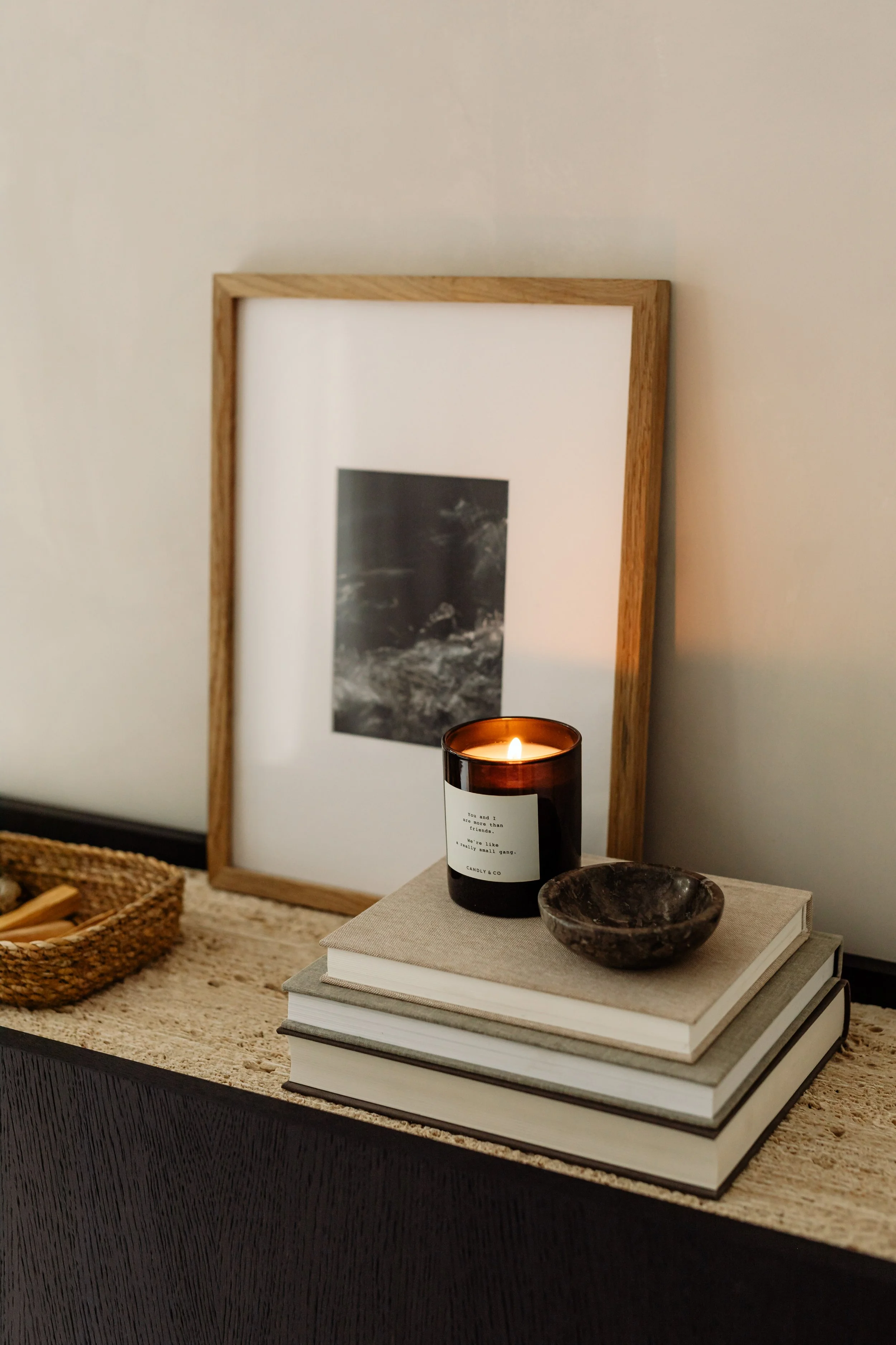 A cozy home decor arrangement featuring a framed black-and-white photograph on a wall, a lit candle on a stack of books, a marble bowl, and a woven basket on a textured surface.