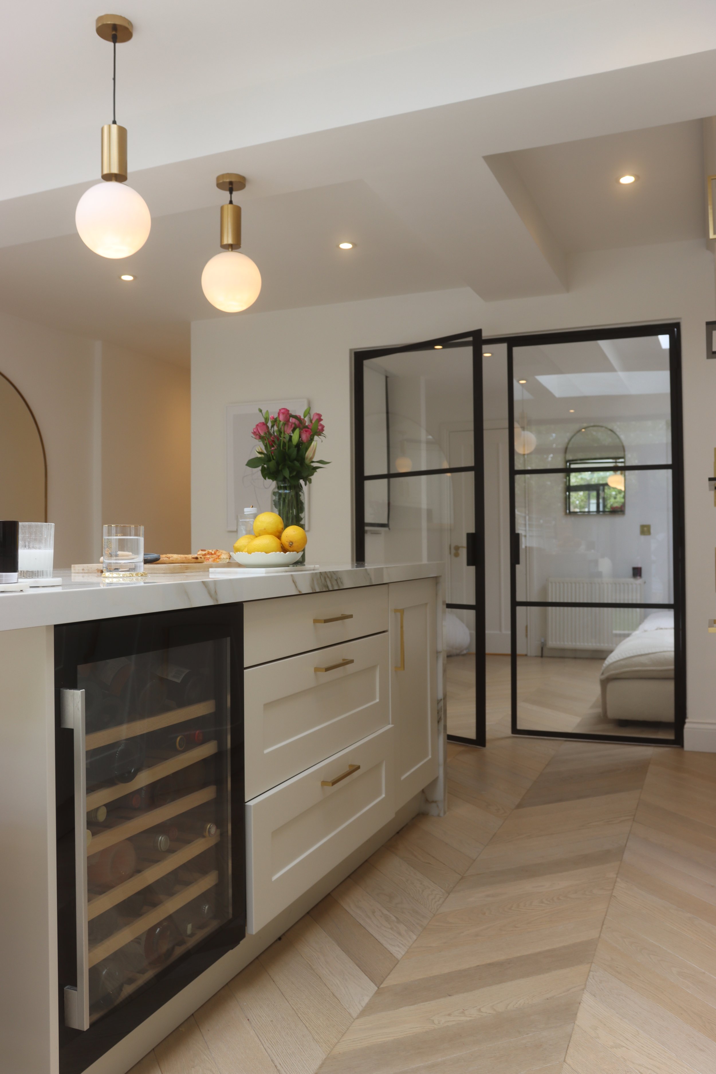 Modern kitchen with pendant lights, a marble countertop, a bowl of lemons, a wine fridge, and a glass door leading to a living area with a sofa and a window.