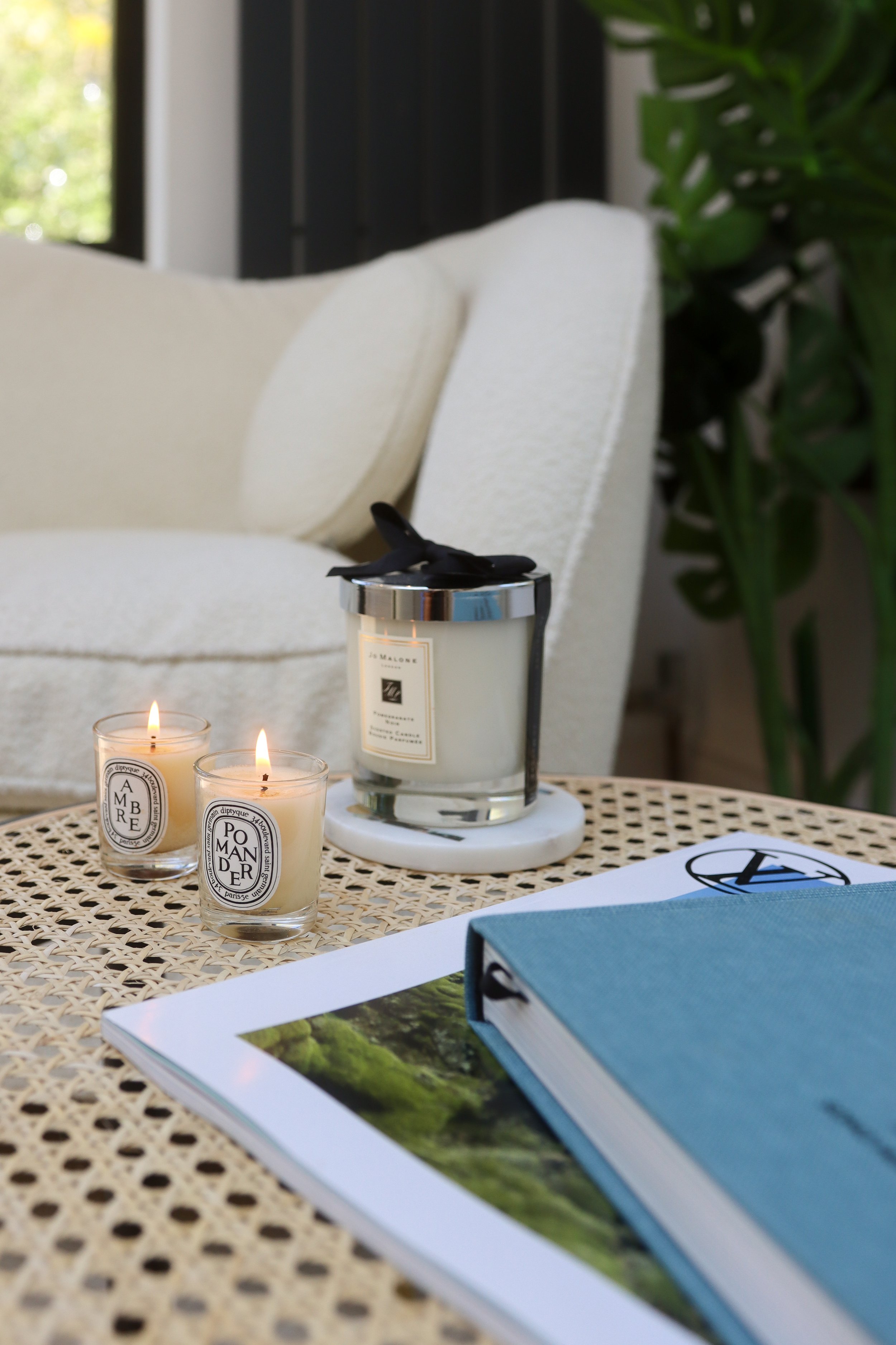 A table with three small lit candles, a blue book on a black and woven surface, with a white sofa, green plant, and window in the background.
