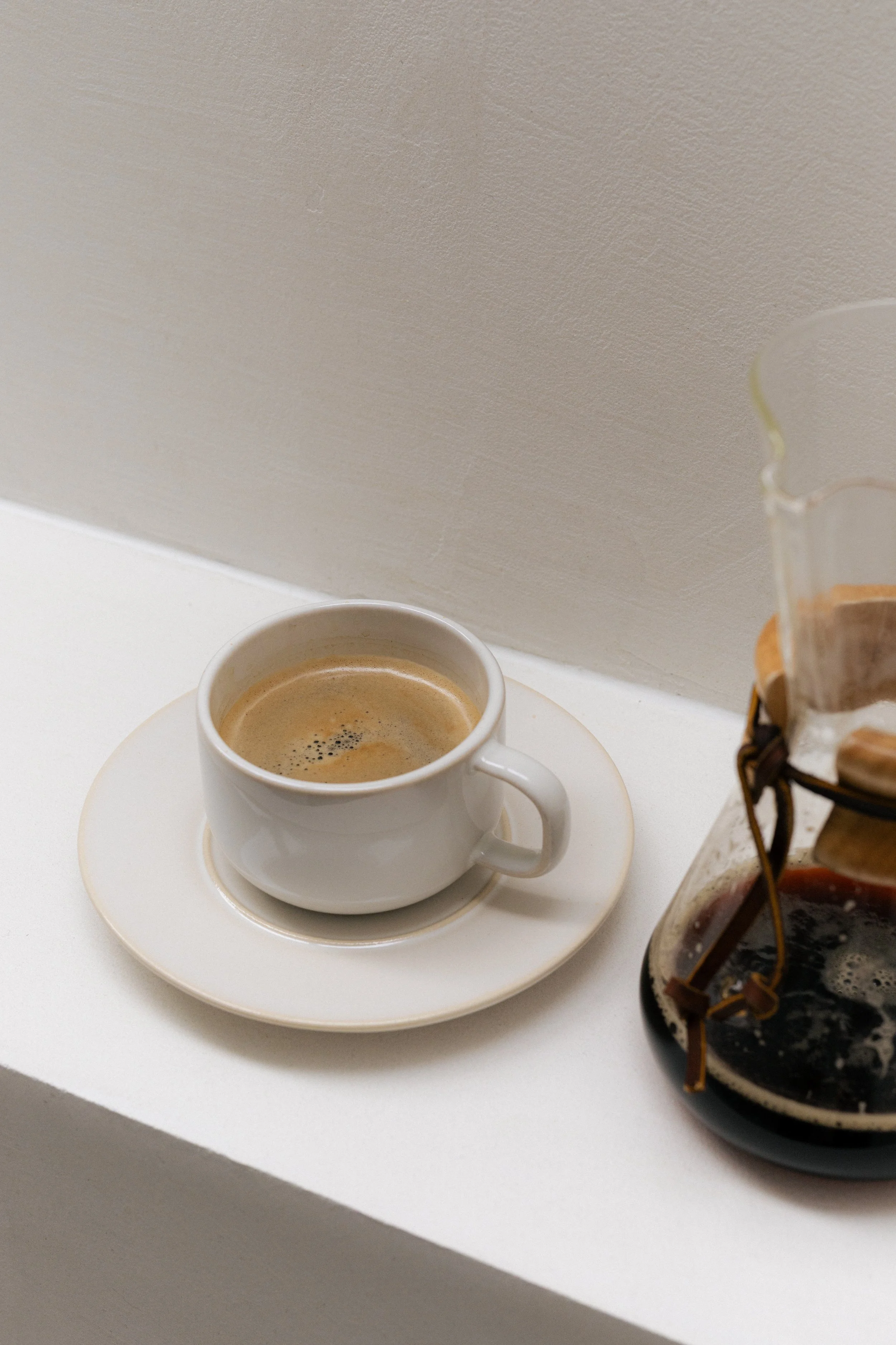 A white coffee cup filled with black coffee on a white saucer, placed on a white surface. Part of a glass jar with a dark substance and metal clips is visible on the right side of the image, against a plain beige wall background.