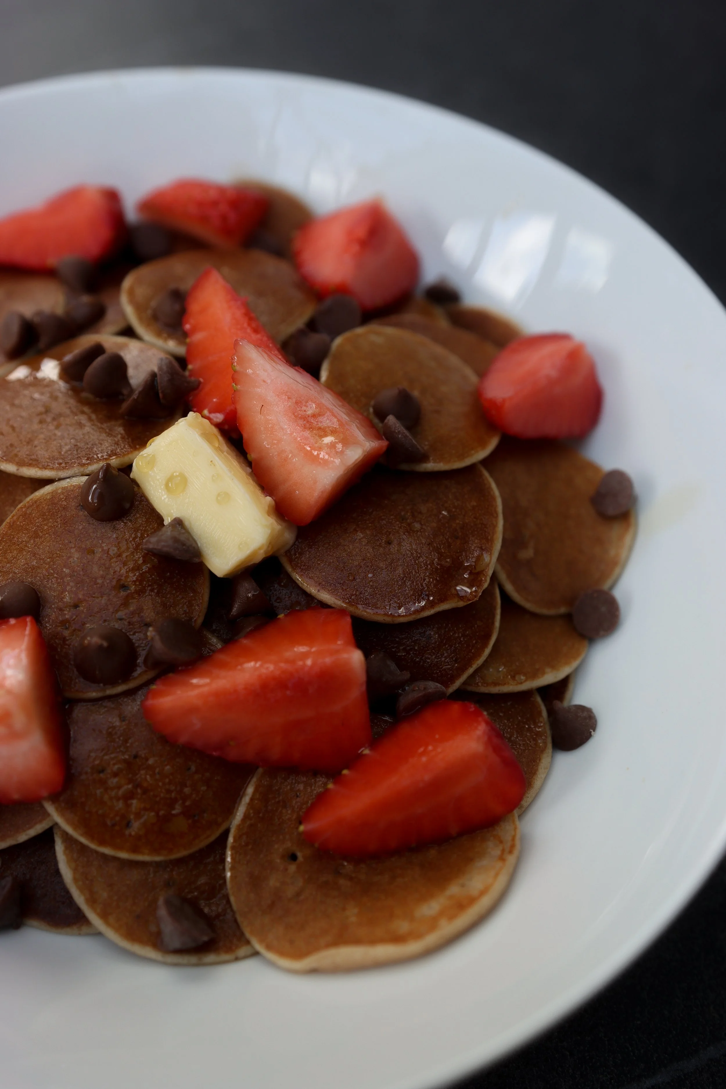 Plate of small pancakes topped with sliced strawberries, chocolate chips, and a piece of white chocolate.