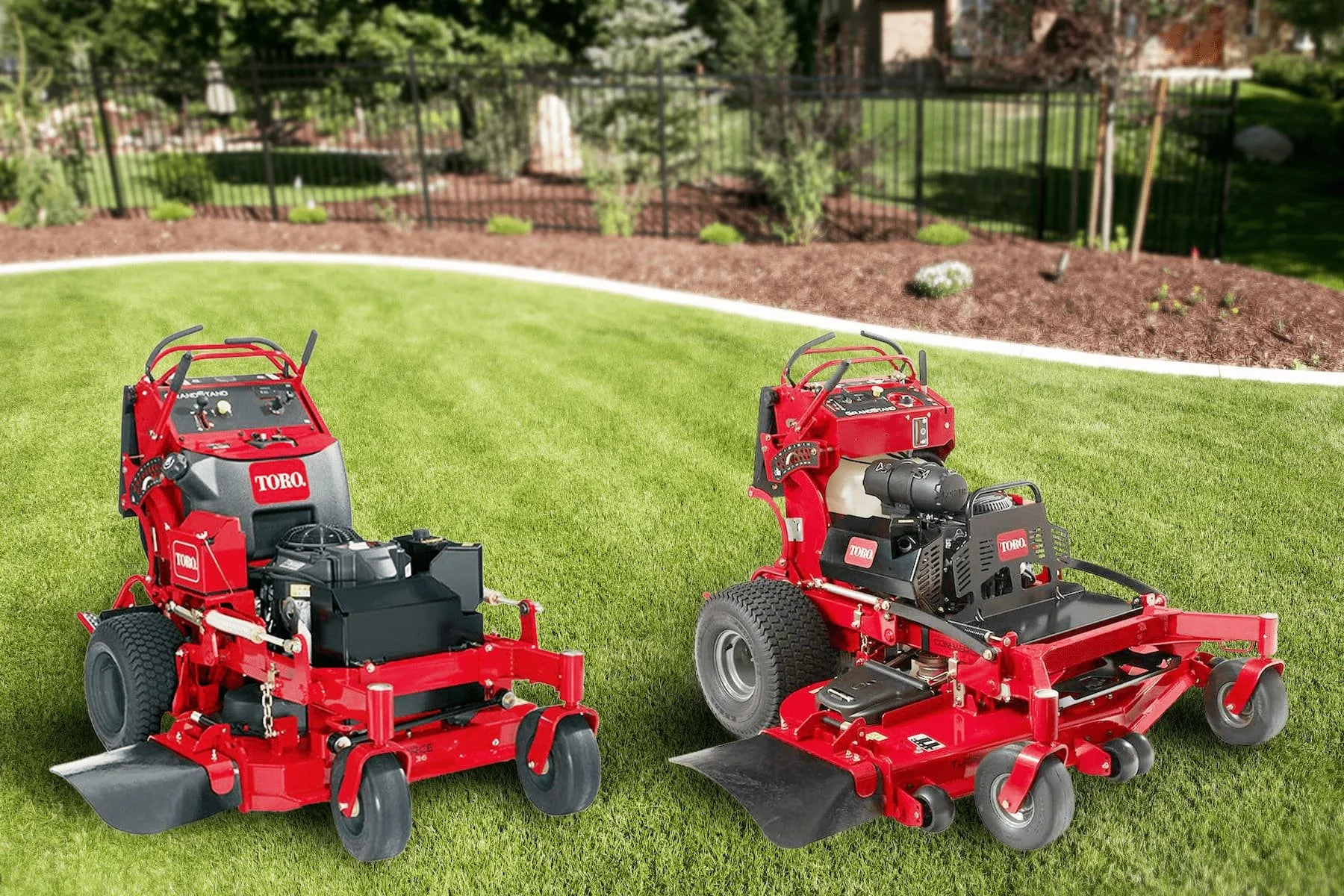 Two red Toro commercial landscaping mowers on a well-maintained green lawn in a residential backyard, with a garden bed and a black fence in the background.