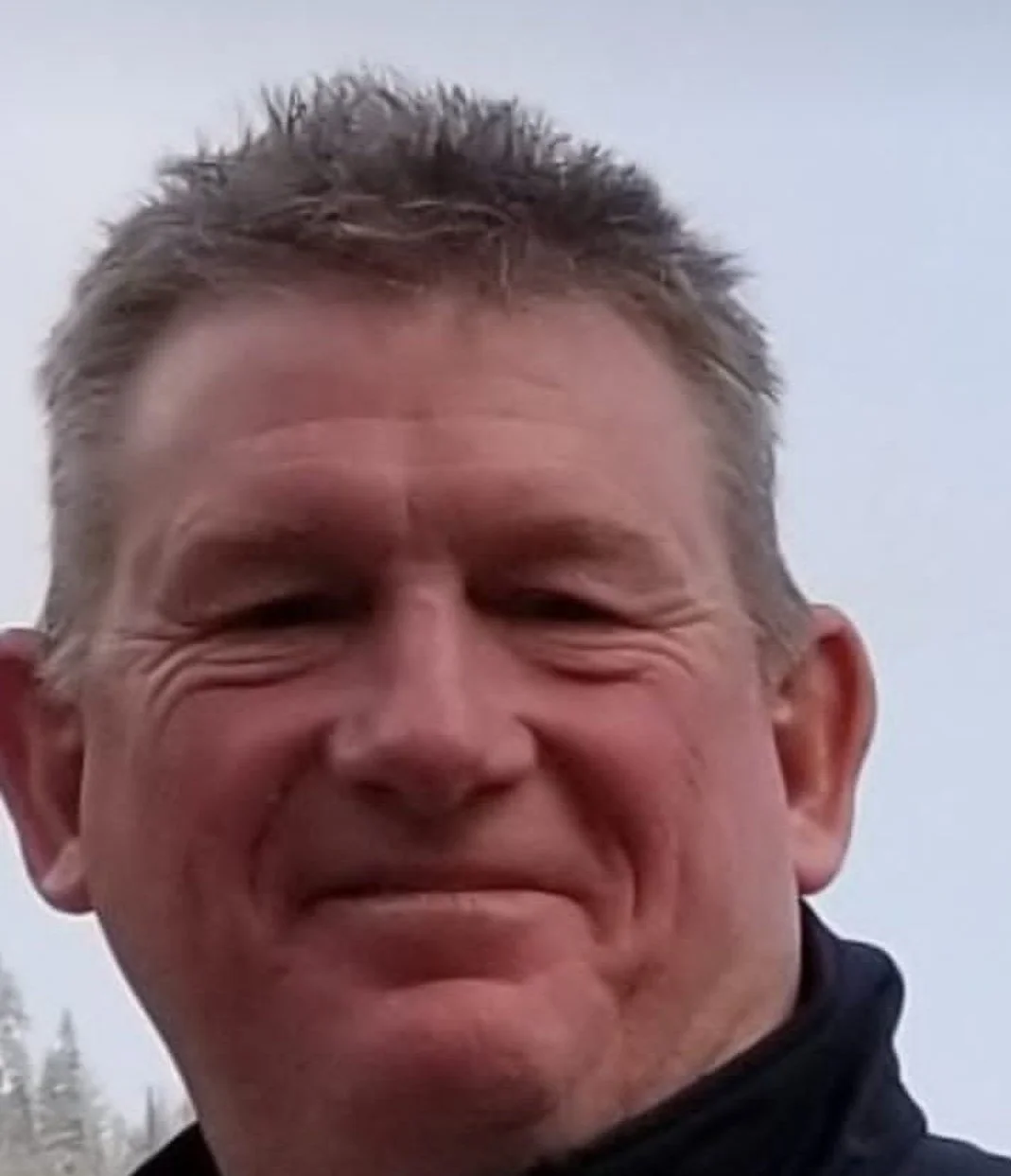 Close-up of a smiling man with short, spiky, light brown hair, outdoors with a cloudy sky in the background.