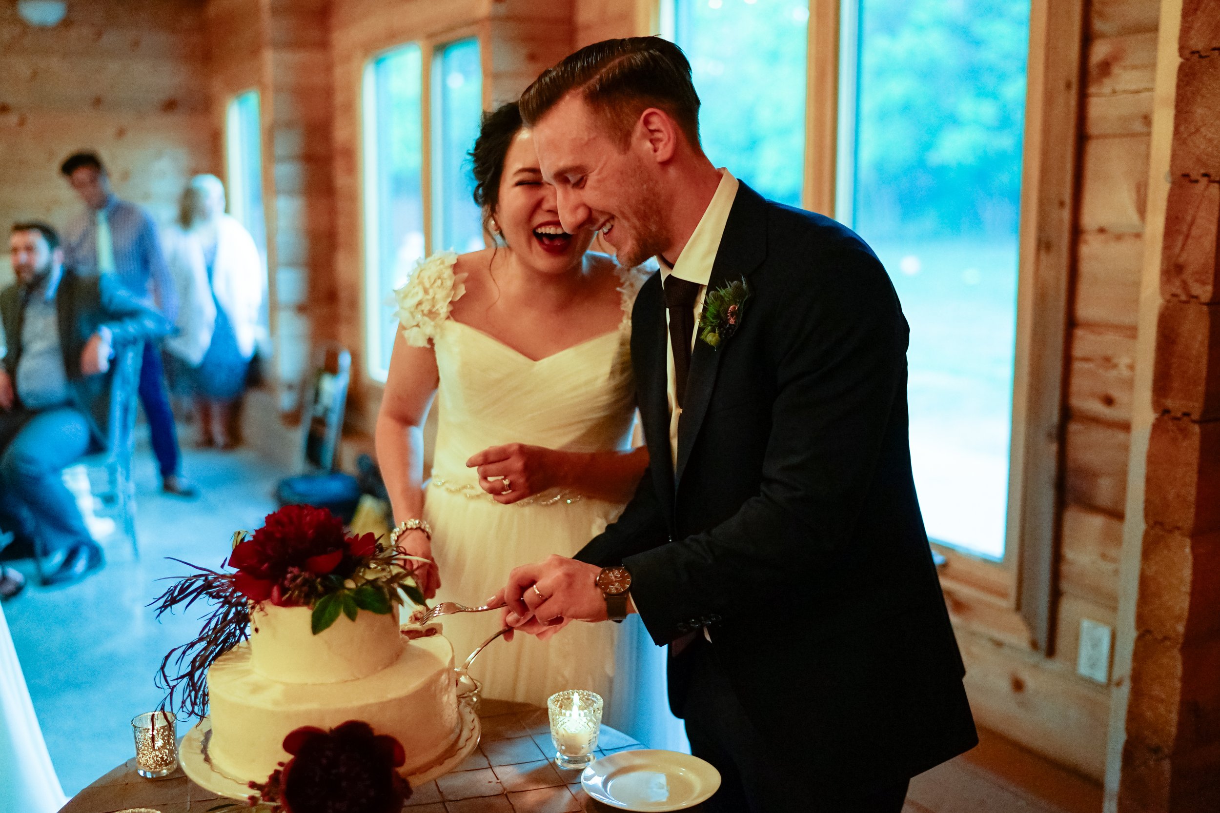 A newlywed couple cuts a wedding cake together in a rustic wooden venue, surrounded by family and friends.