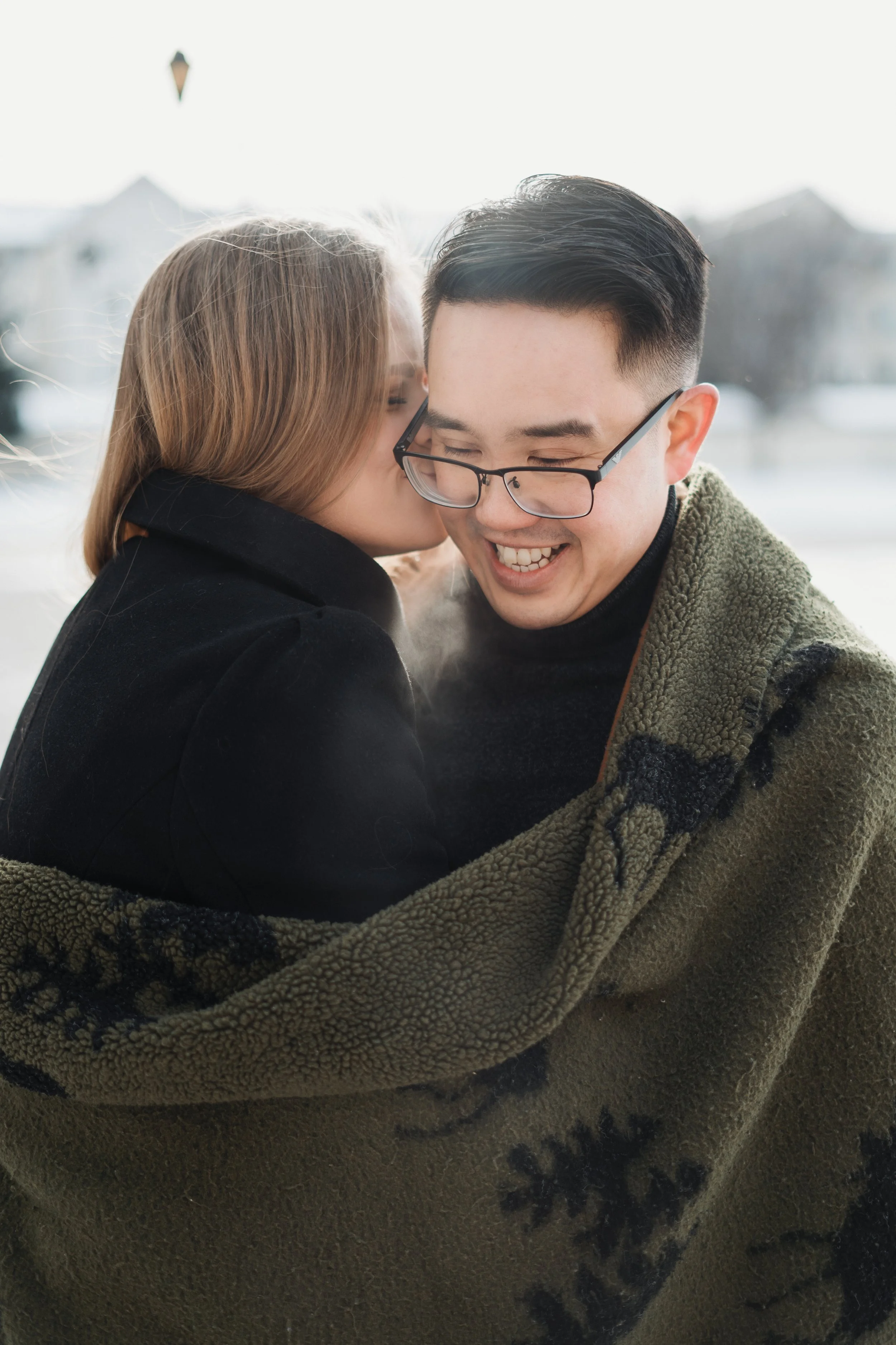 A young woman kisses a smiling young man on the cheek while wrapped in a blanket outdoors on a cold day.