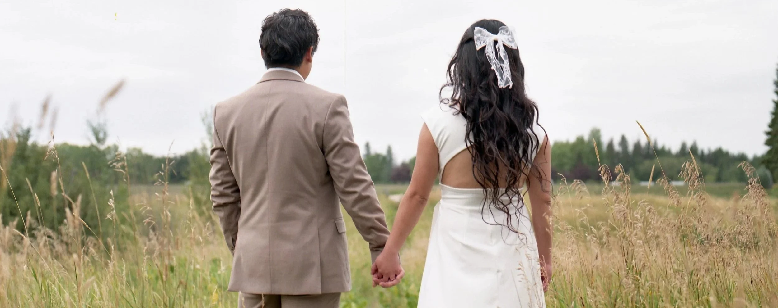 A couple dressed in wedding attire holding hands while walking through a grassy field, with trees in the background and an overcast sky.